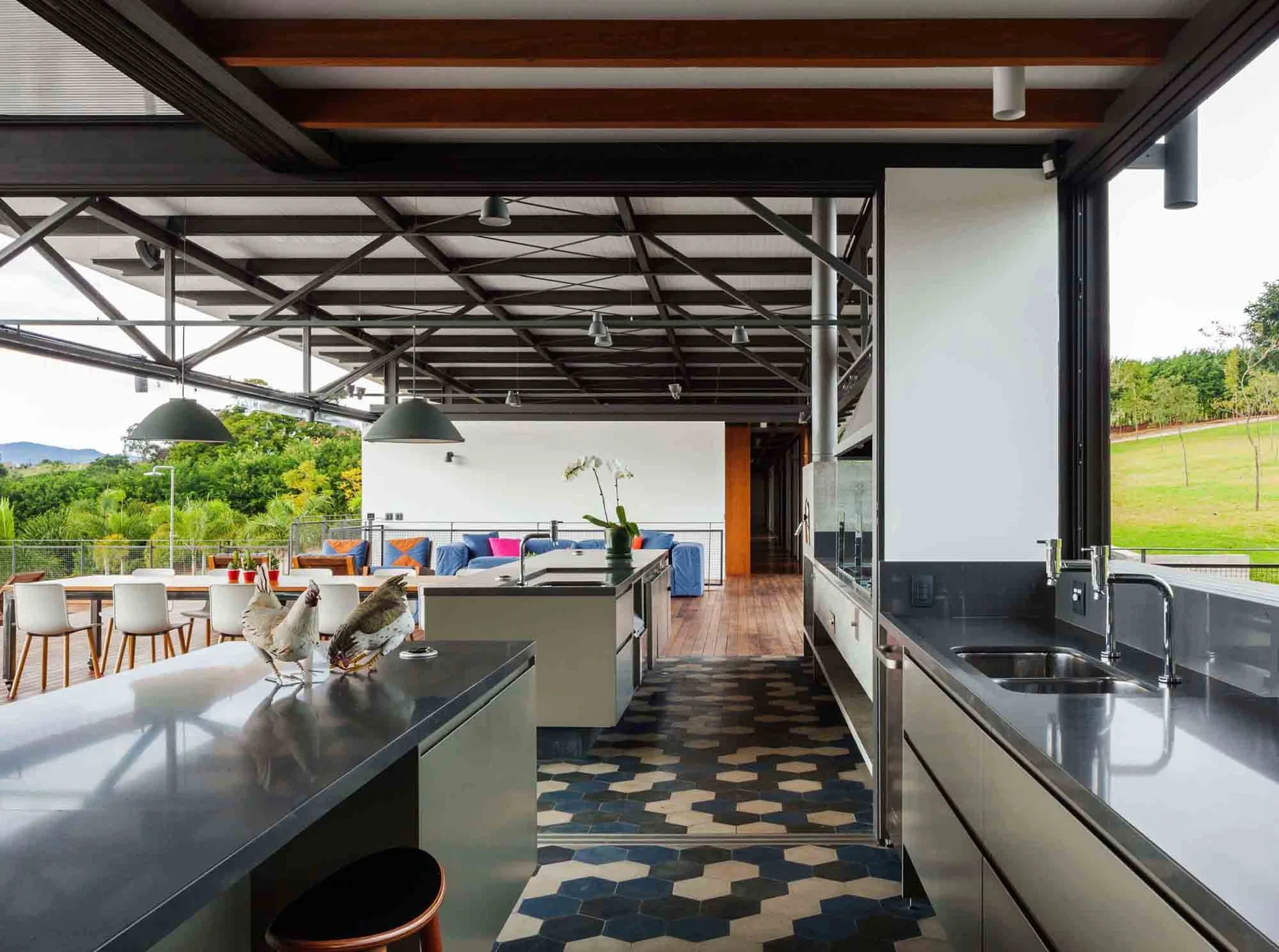 An industrial kitchen with grey cabinetry, a blue and white hexagonal tiled floor, and an exposed steel truss roof that opens to an outdoor terrace.