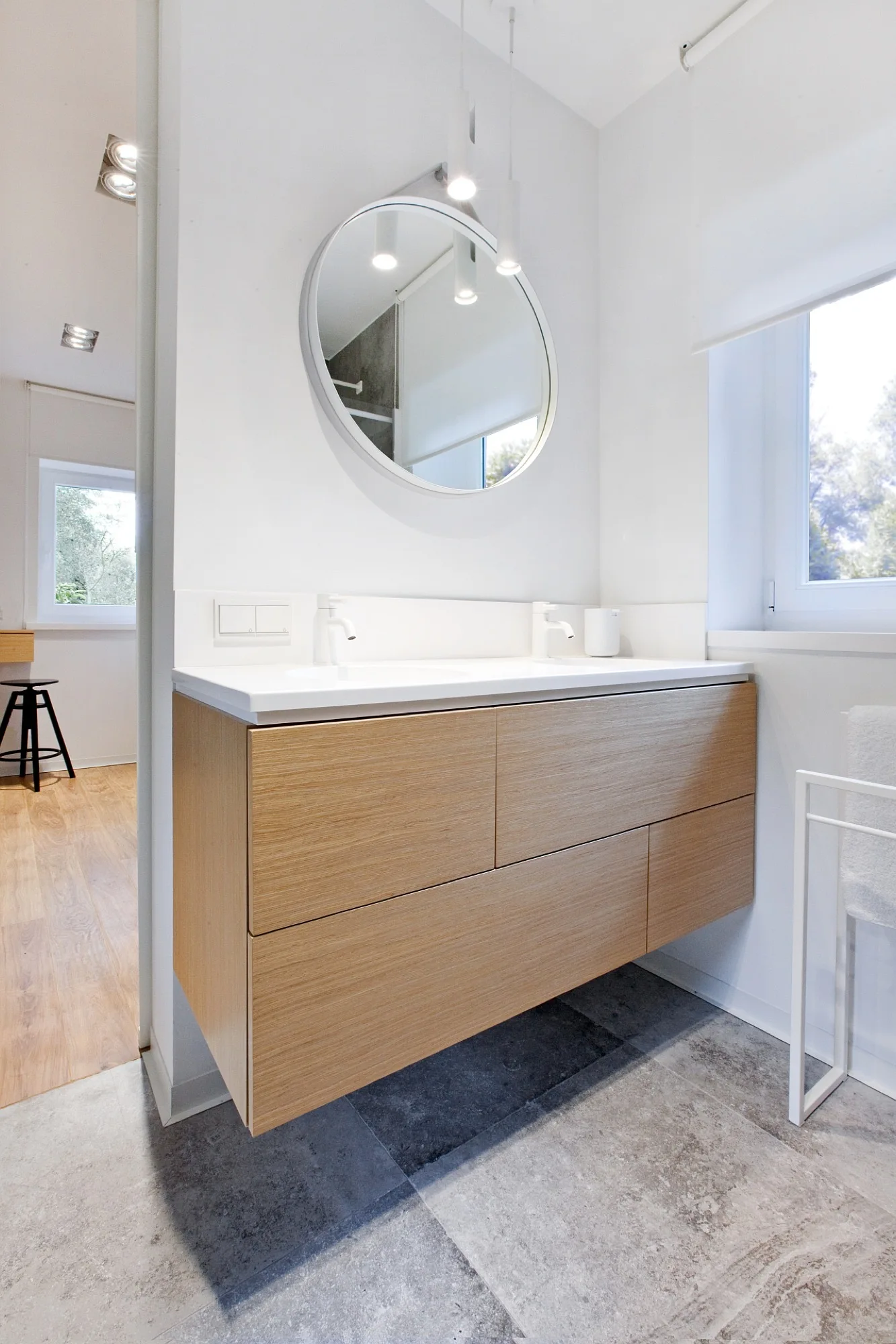 A modern bathroom features a wall-mounted light oak vanity with double sinks, a round white-framed mirror, and grey stone floor tiles.