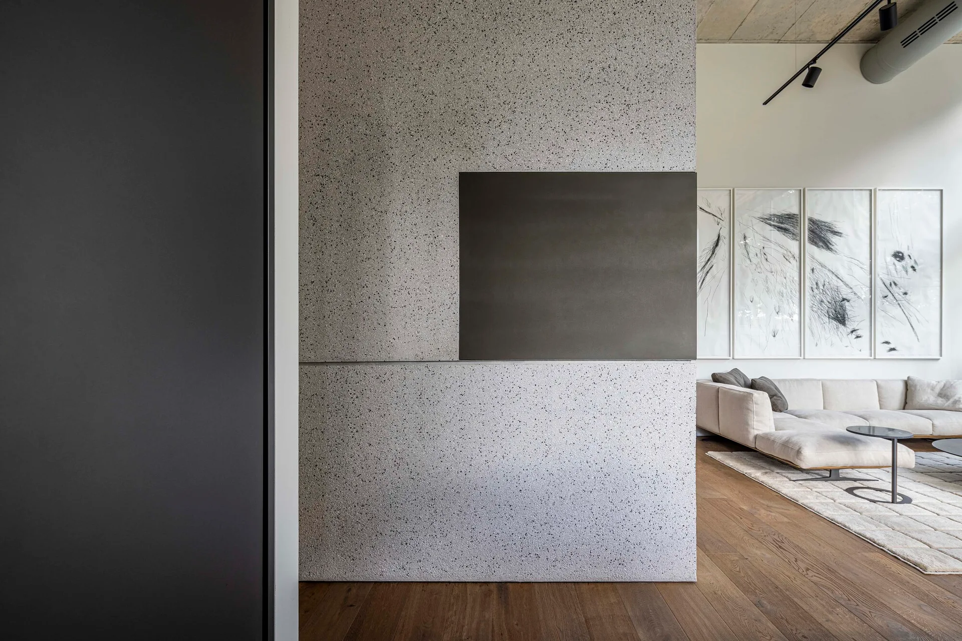 A speckled grey partition wall with a dark rectangular insert stands in front of a modern living room with a beige sectional and wood floors.