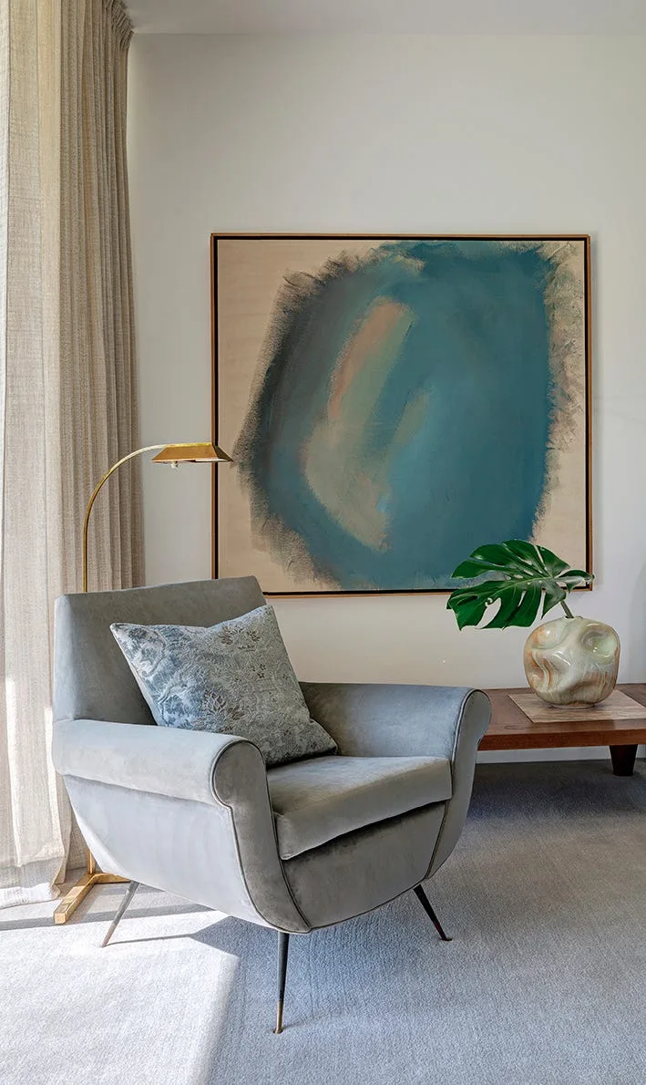 A cozy modern reading corner features a gray upholstered armchair positioned in front of a large teal abstract painting.