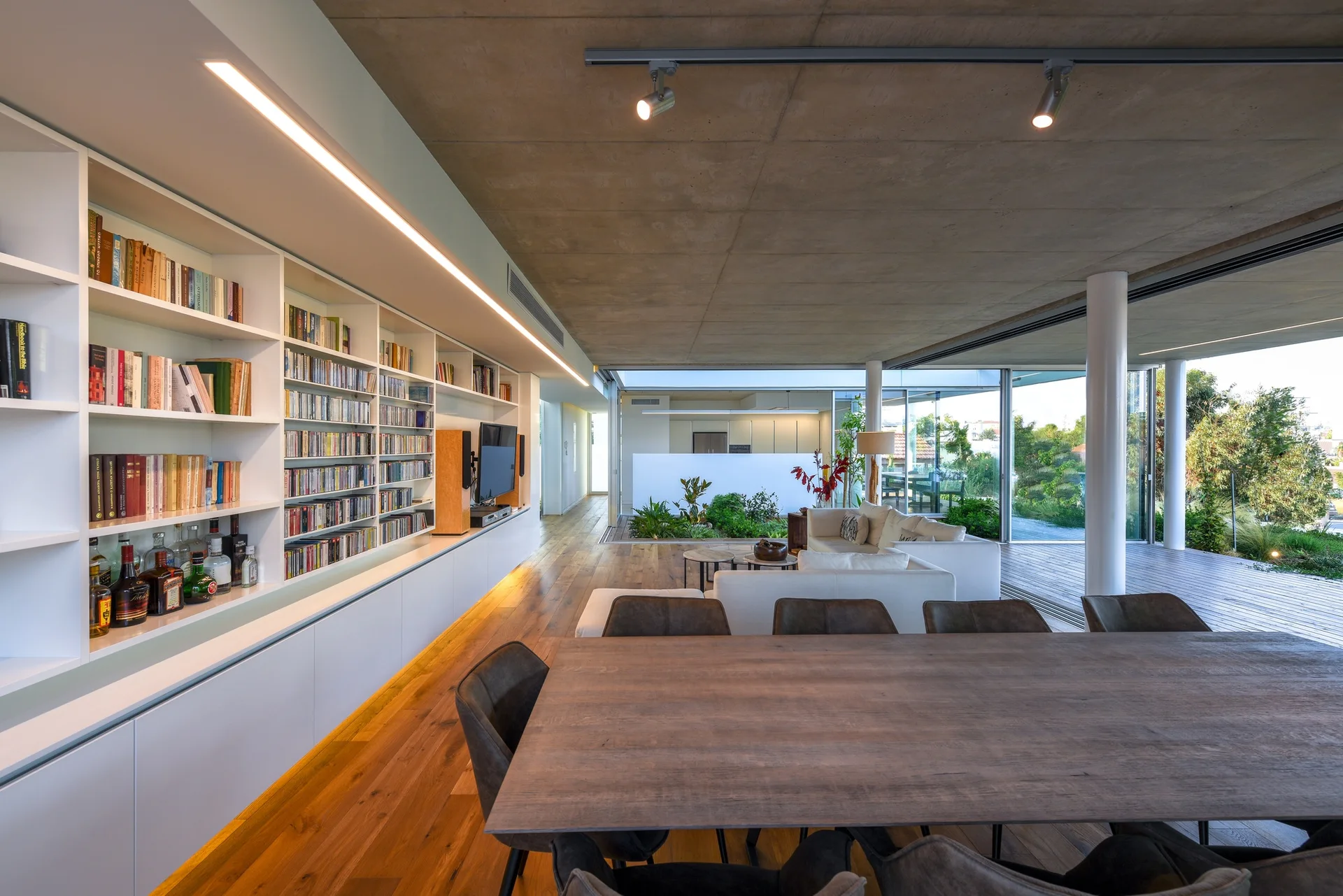 An open-plan living and dining area featuring an exposed concrete ceiling, a long built-in bookshelf wall, a dining table, and large glass windows.
