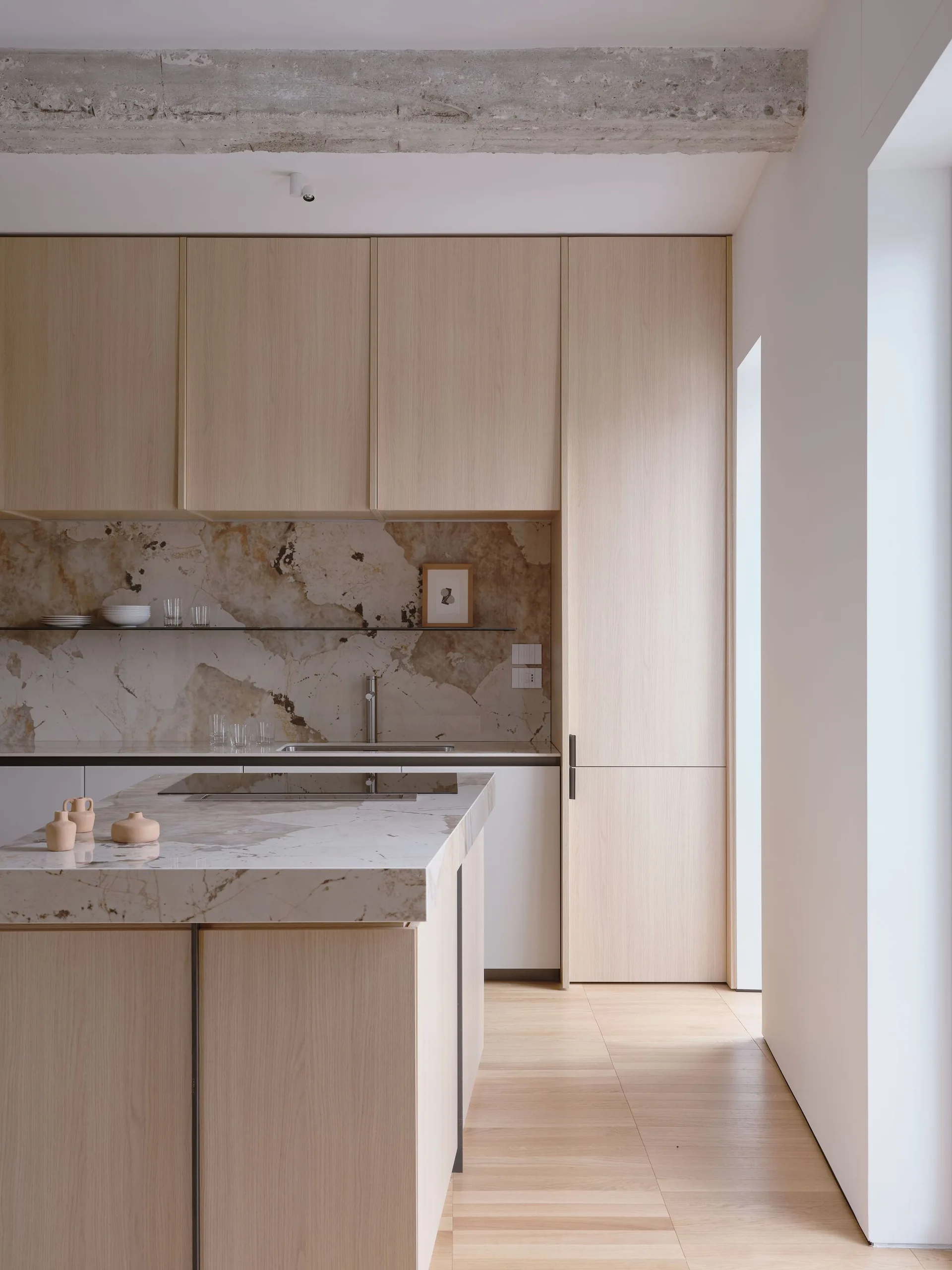 A minimalist kitchen features light oak flat-front cabinets, a highly veined stone slab backsplash and island, and an exposed concrete ceiling beam.