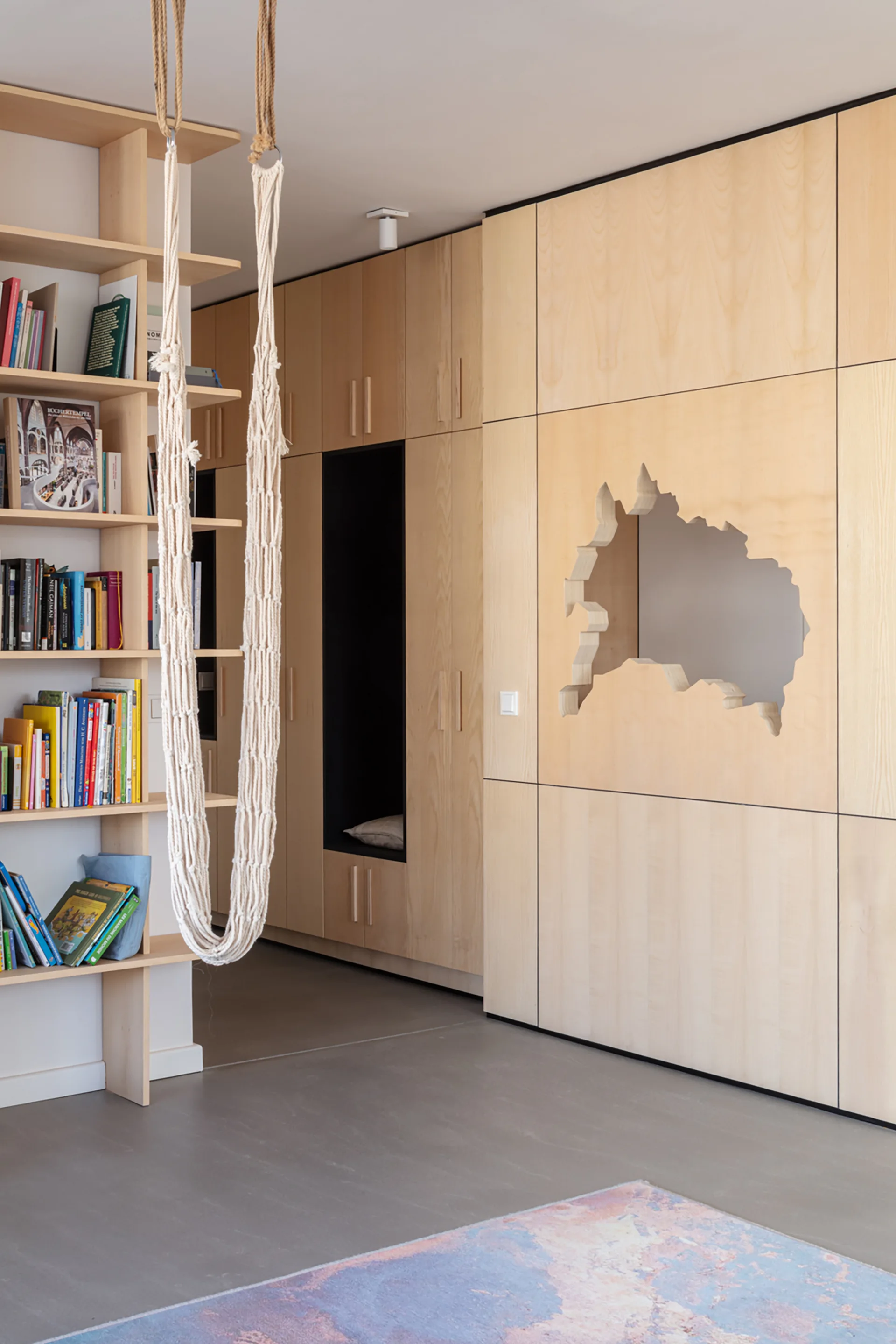 A light wood built-in wardrobe features an abstract cutout alongside an open bookshelf and a hanging white macrame swing.