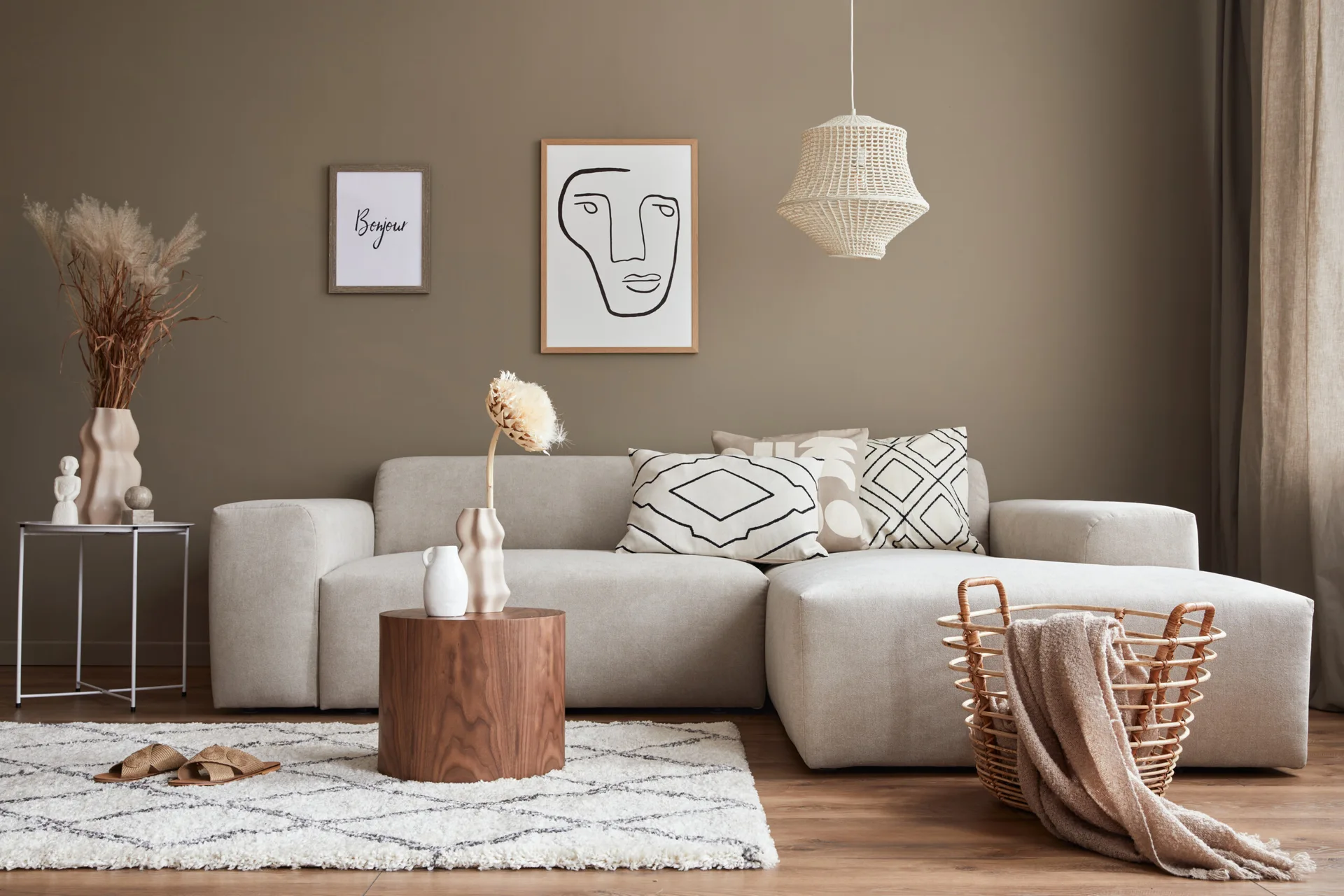 A light cream sectional sofa sits against a taupe wall, paired with a round wooden coffee table, a diamond-patterned shag rug, and a woven pendant light.