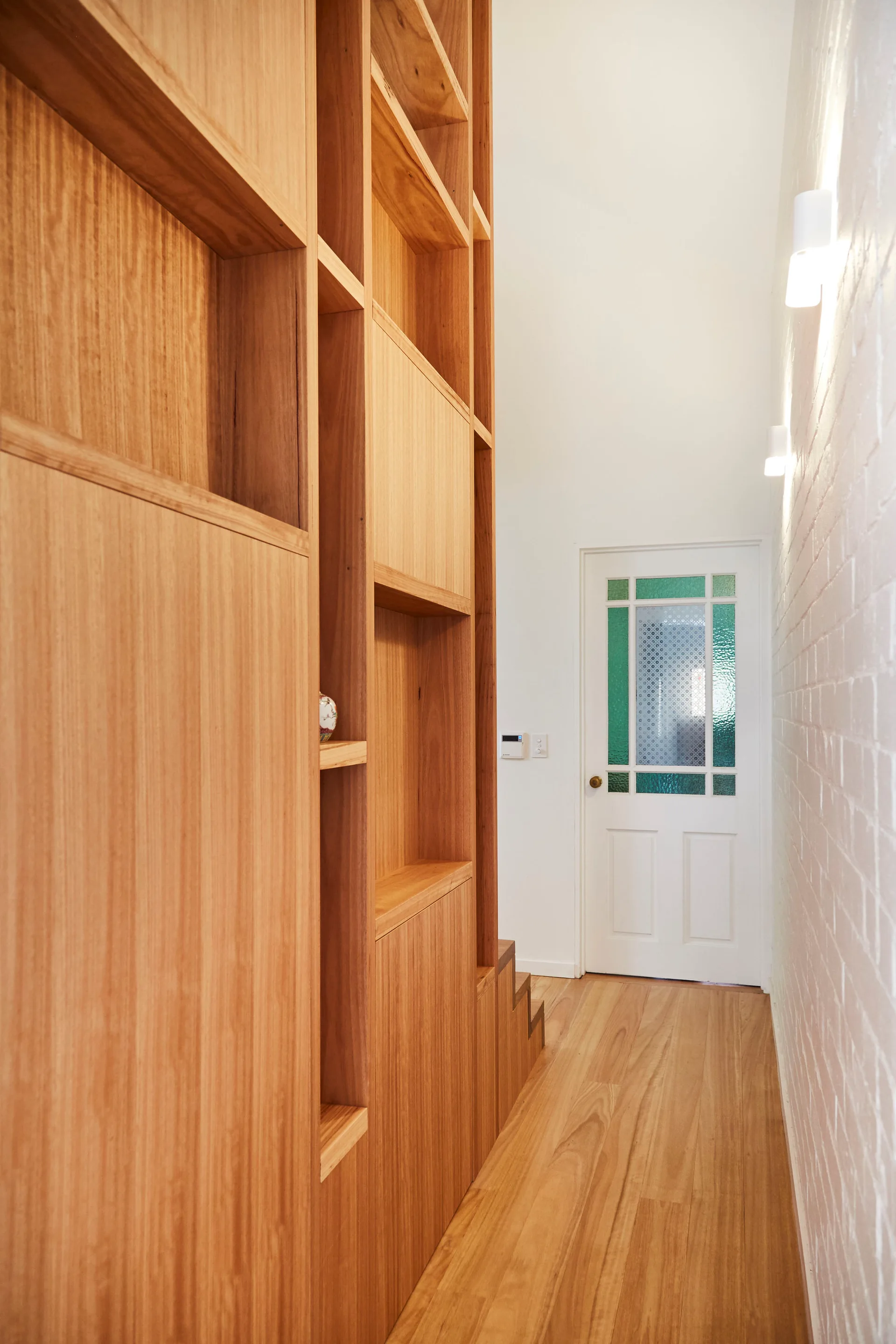 A narrow hallway features a tall wooden built-in cabinet opposite a white painted brick wall with sconces, leading to a door with green glass panels.