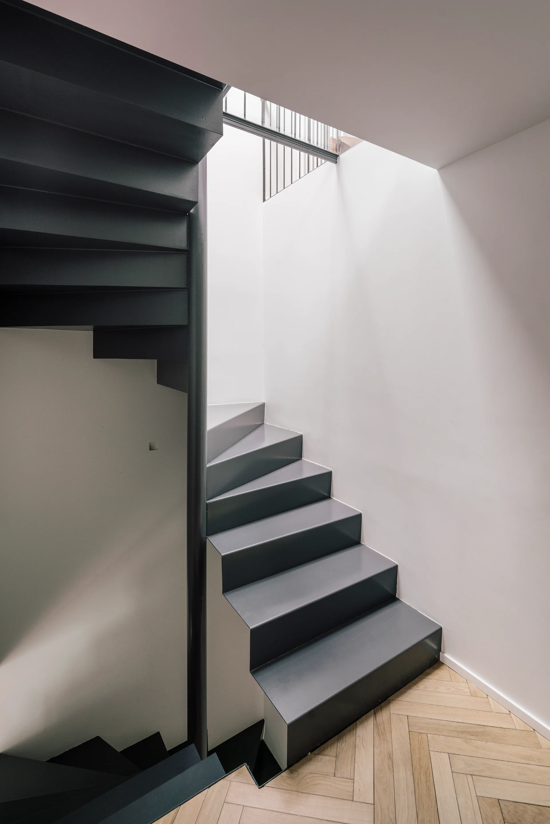 A dark gray steel staircase twists upward alongside clean white walls, landing on light herringbone-patterned wood floors.