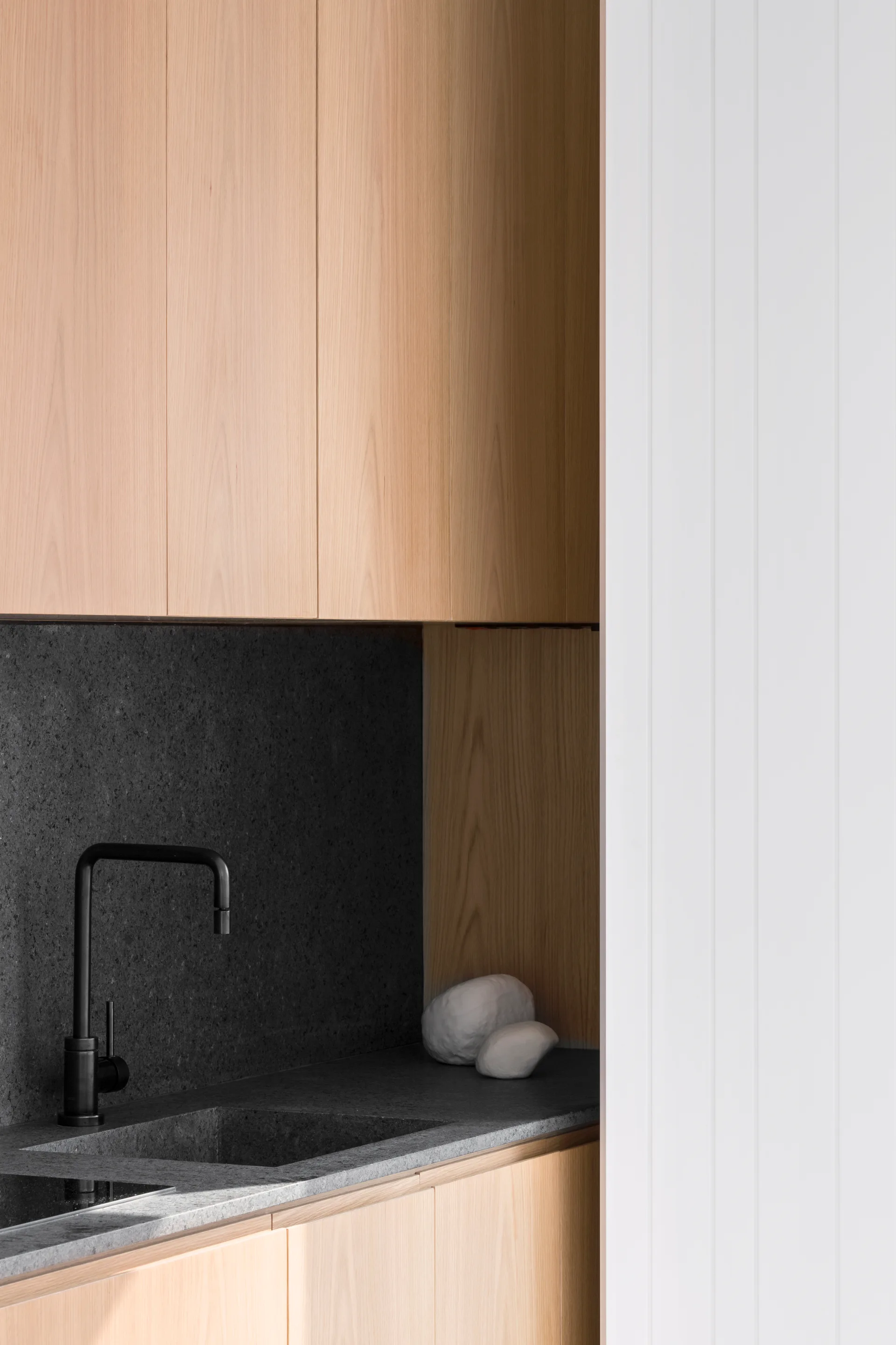 A minimalist vanity features light wood cabinets, a dark grey stone countertop with an integrated sink, a matte black faucet, and white paneled walls.