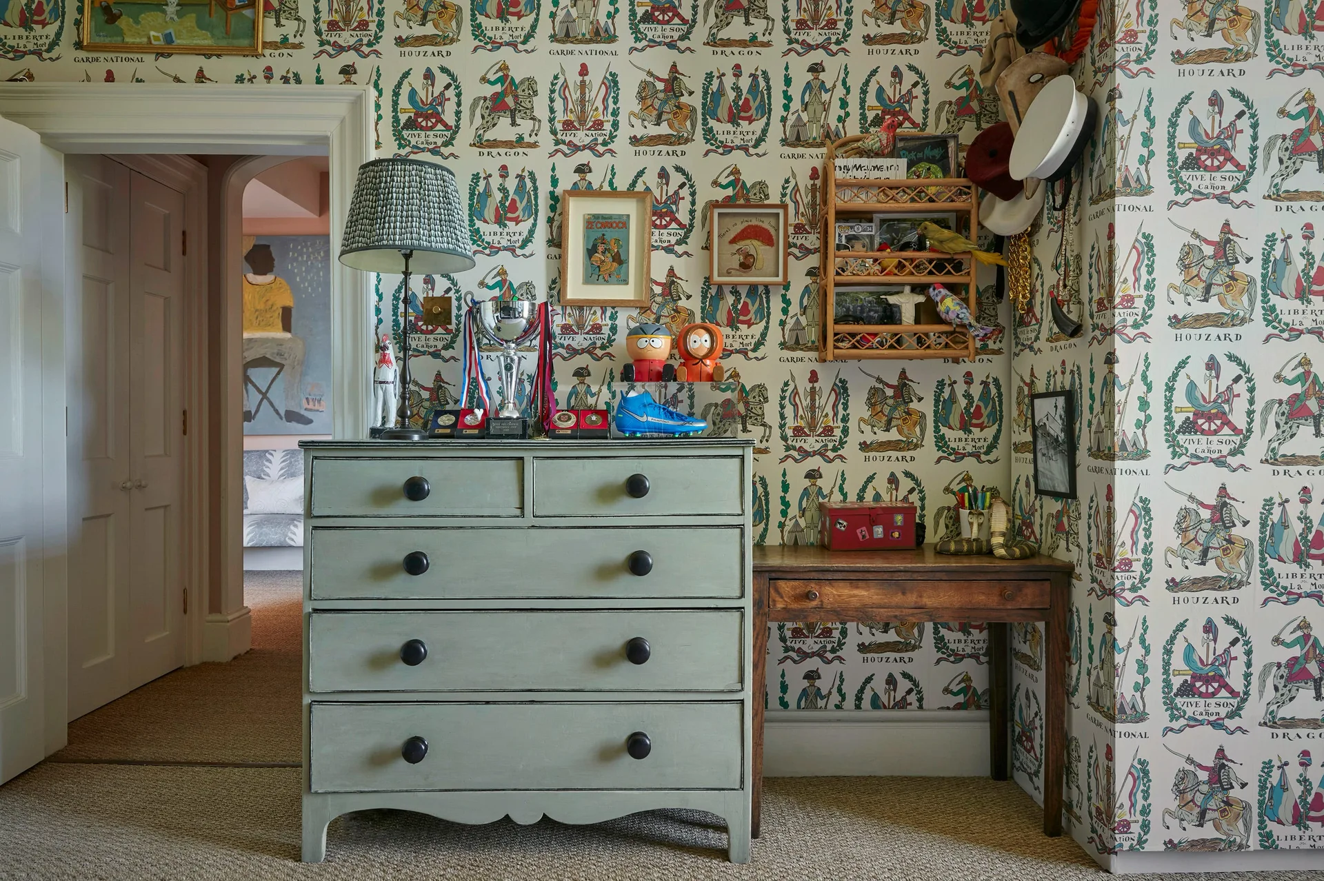A sage green dresser and a small wooden desk stand against a wall covered in illustrative military-themed wallpaper in a bedroom corner.