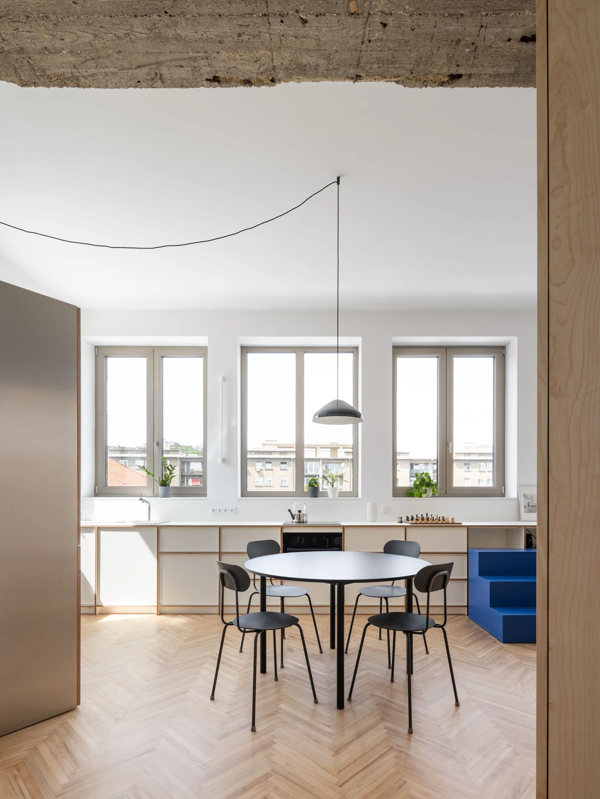A minimalist dining room features a round black table, matching chairs, herringbone wood floors, and low white cabinets under three large windows.