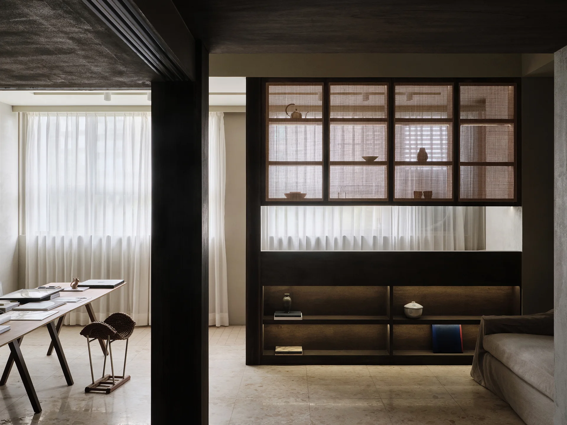 A minimalist home office featuring a wooden desk, a woven stool, sheer white curtains, and dark built-in shelves with woven panels.