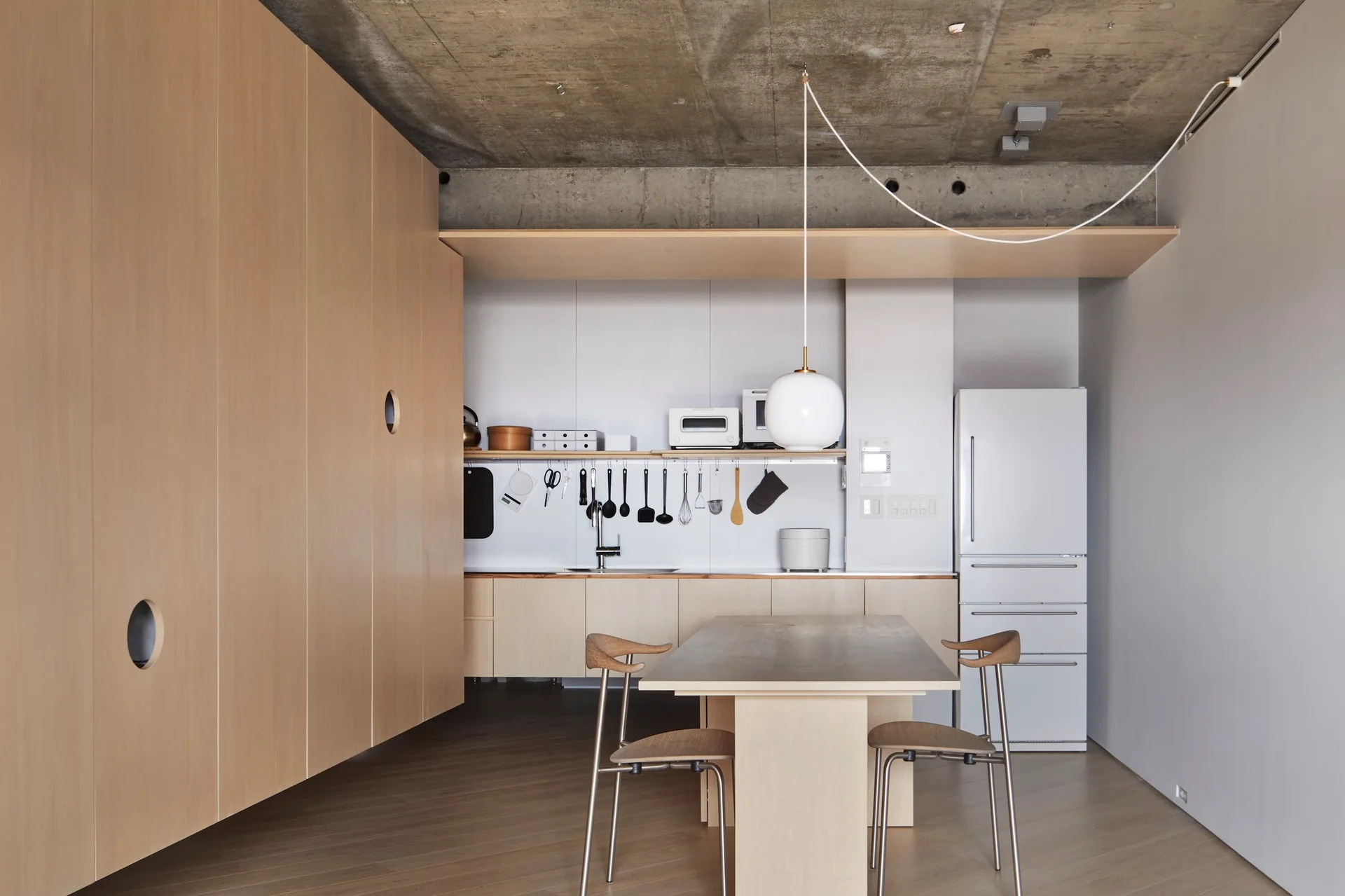 A minimalist kitchen featuring floor-to-ceiling light wood cabinets, an exposed concrete ceiling, and a simple dining table set.