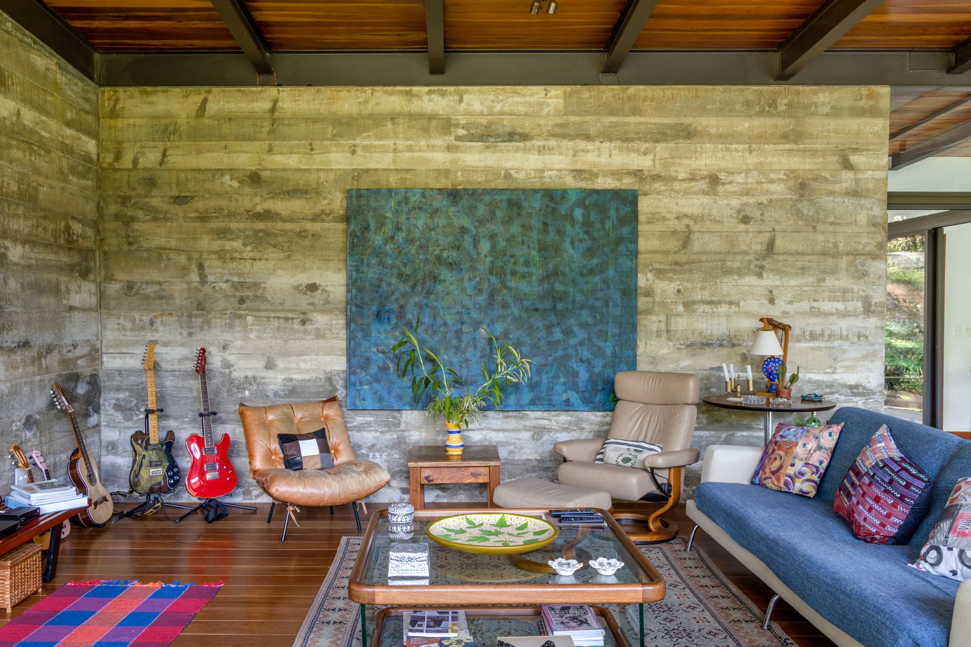 An eclectic living room displays a blue sofa, leather armchairs, and guitars resting against a textured concrete wall under a wood ceiling.