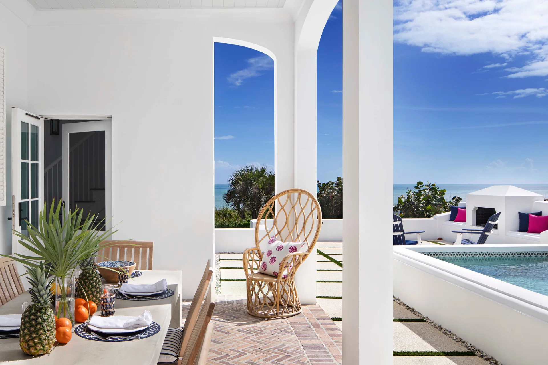 A white arched patio features a dining table, a high-back rattan chair on a herringbone brick floor, and a swimming pool overlooking the ocean.