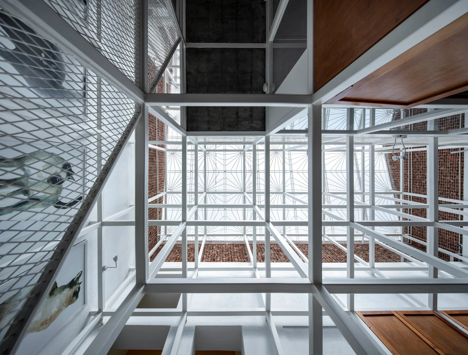 A low-angle view looking up through a multi-story atrium with a white metal grid framework, exposed brick walls, and a glass skylight ceiling.
