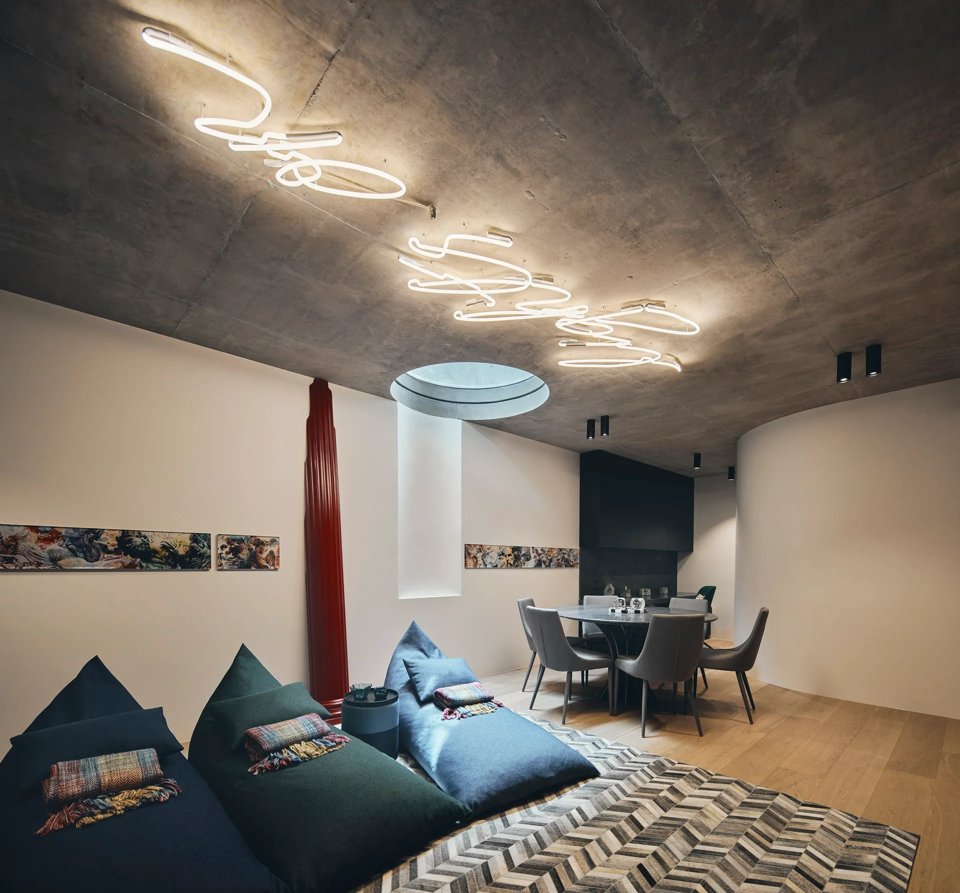 An open-plan living room features three bean bag chairs on a geometric rug beneath an industrial concrete ceiling with a custom neon light fixture.