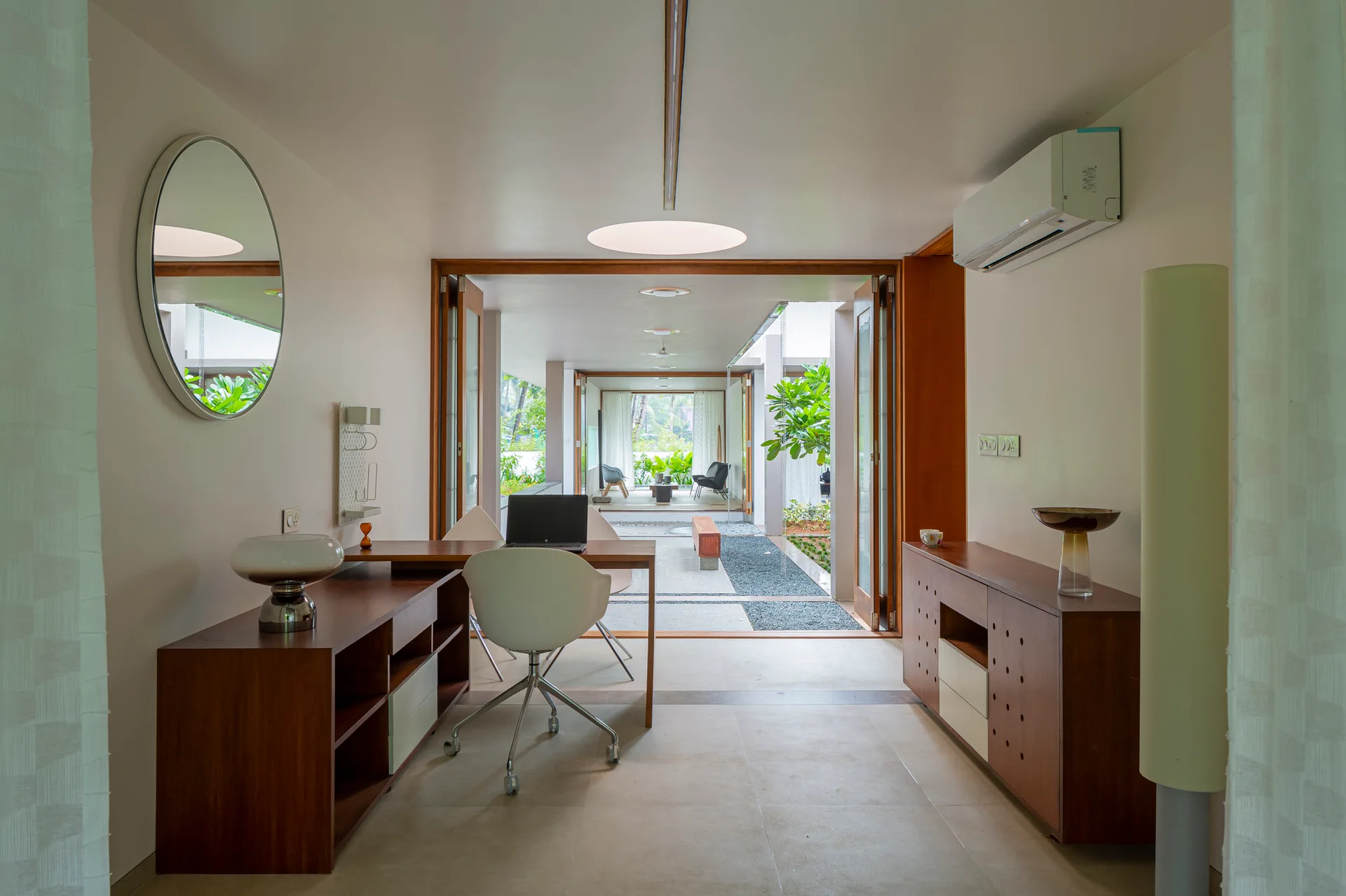 A minimalist home office features a wooden desk, white office chair, and wooden sideboard, opening directly onto a gravel courtyard walkway.