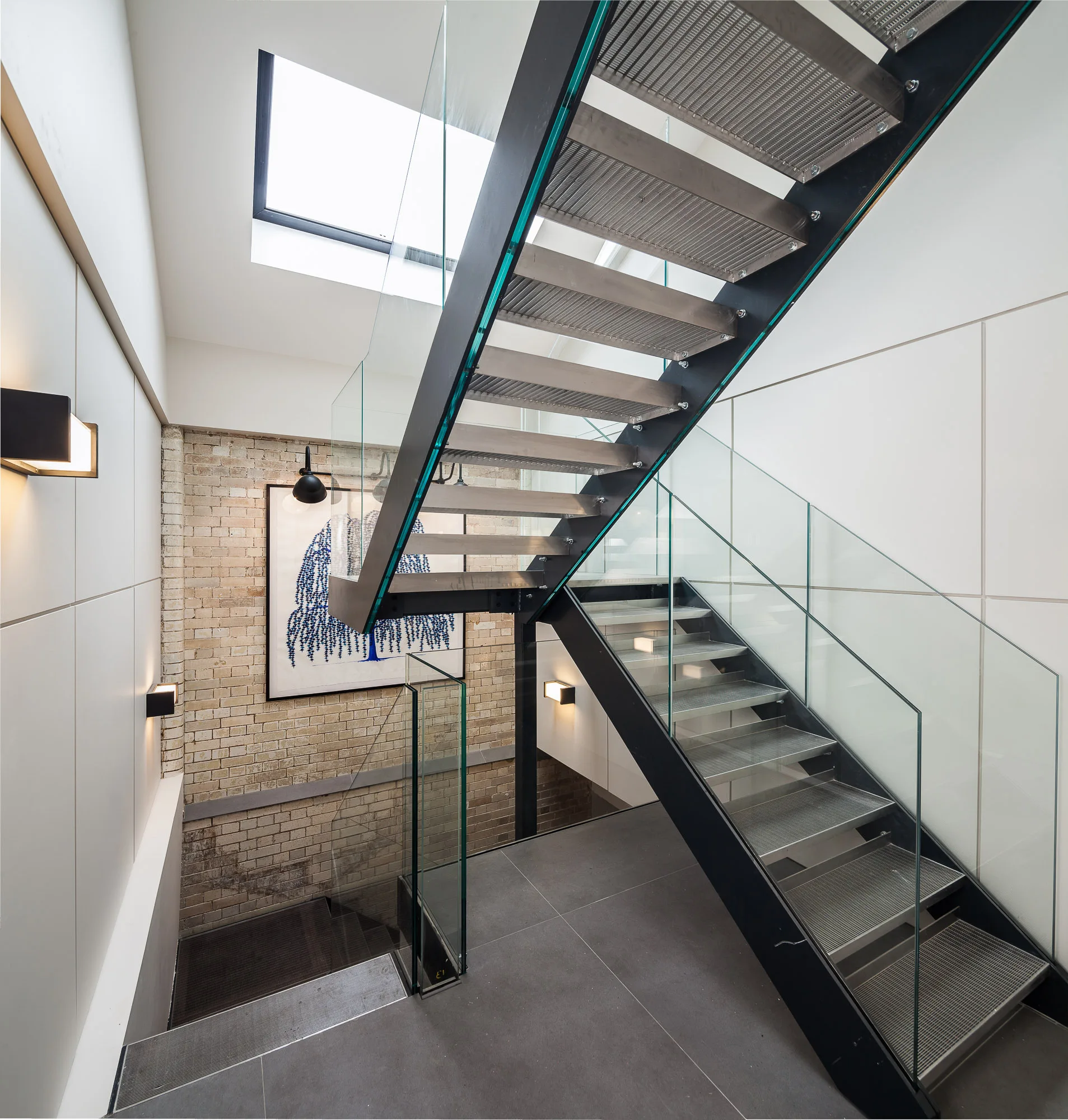 A multi-level metal and glass staircase stands in a bright stairwell featuring an exposed brick wall, a skylight, and grid-patterned wall panels.