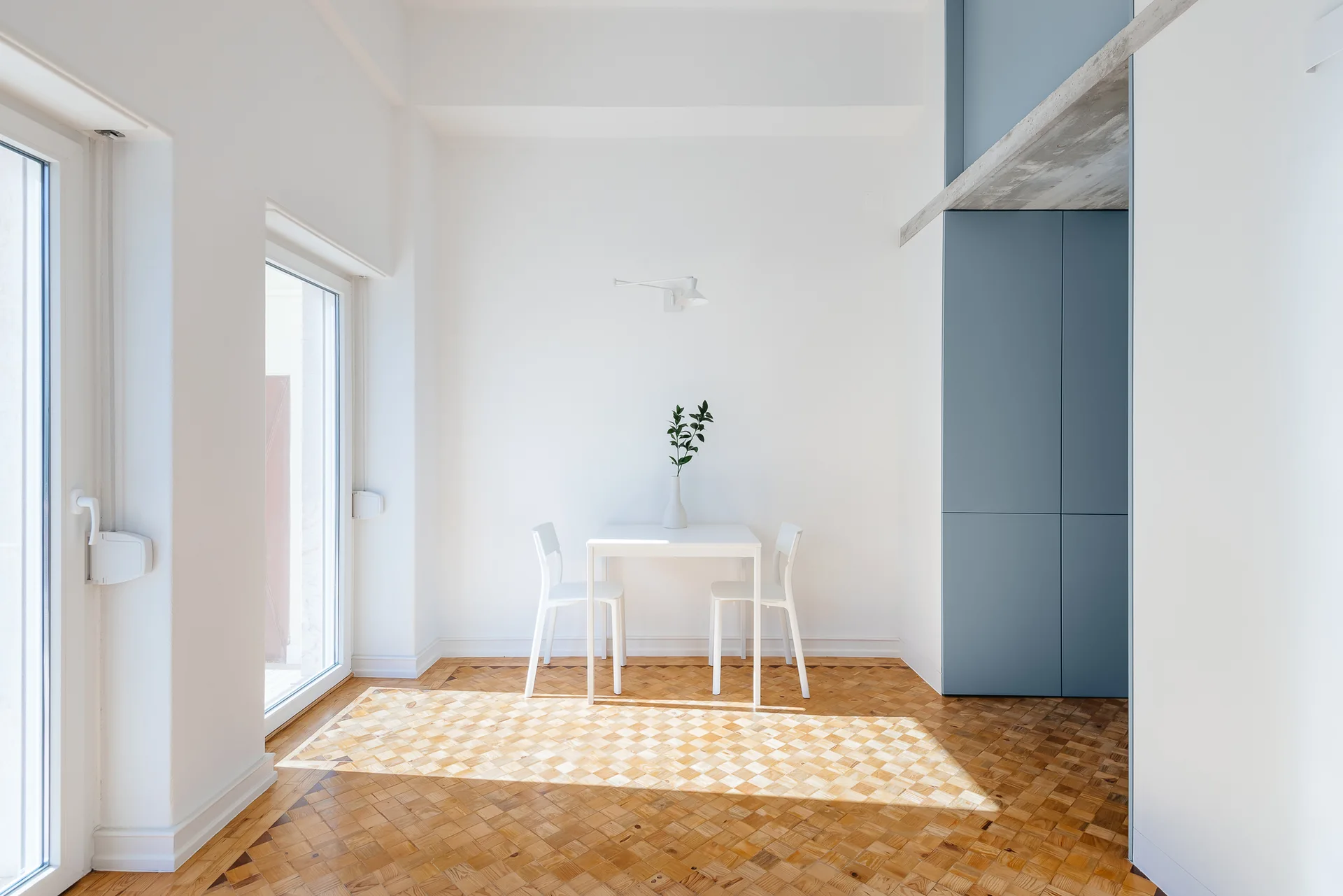 A minimalist white dining table and two chairs stand on a light wood parquet floor next to a tall blue cabinet in a sunlit room.