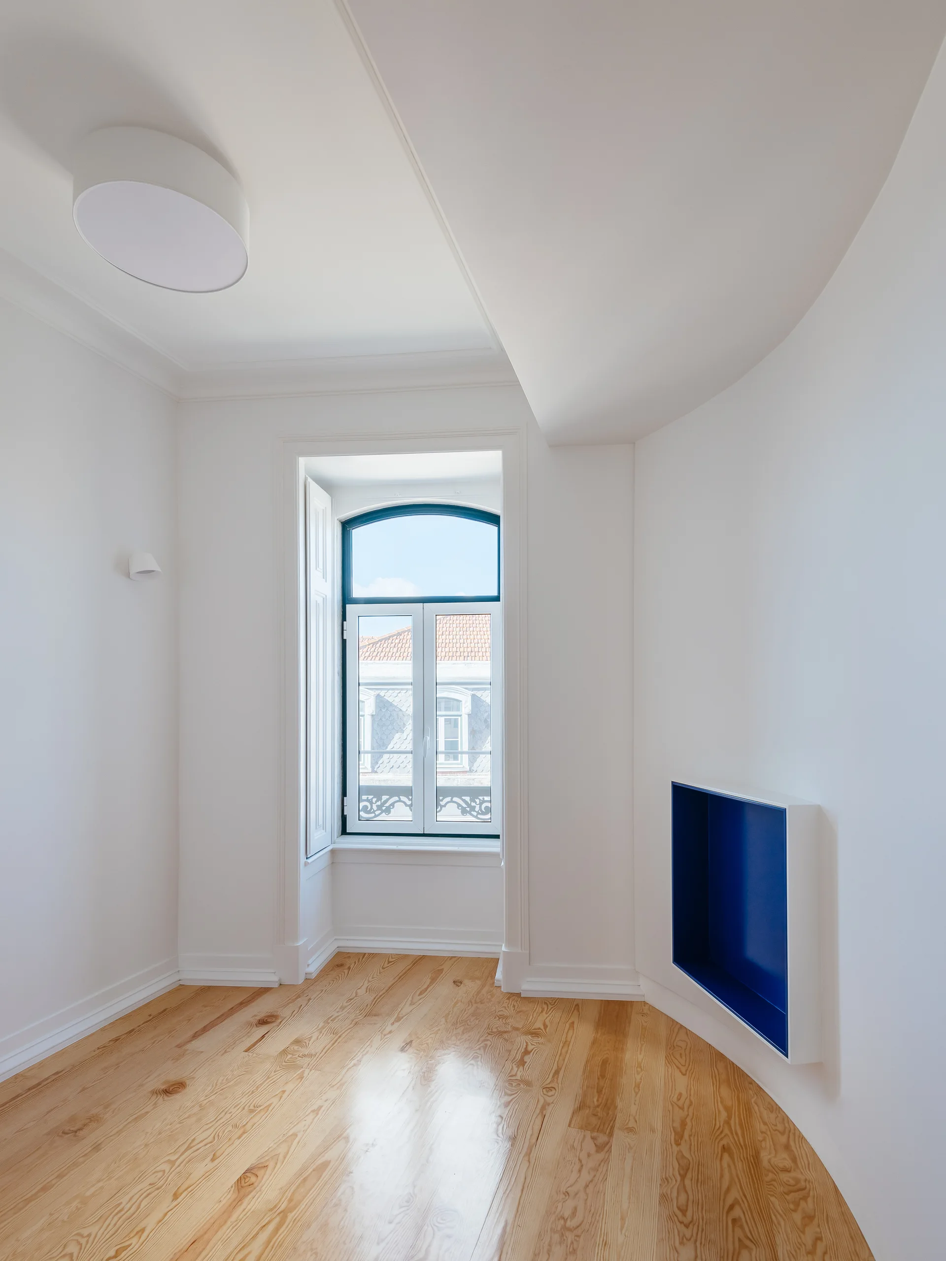 An empty room with polished light wood floors, curved white walls, an arched window, and a recessed rectangular shelf painted bright blue.