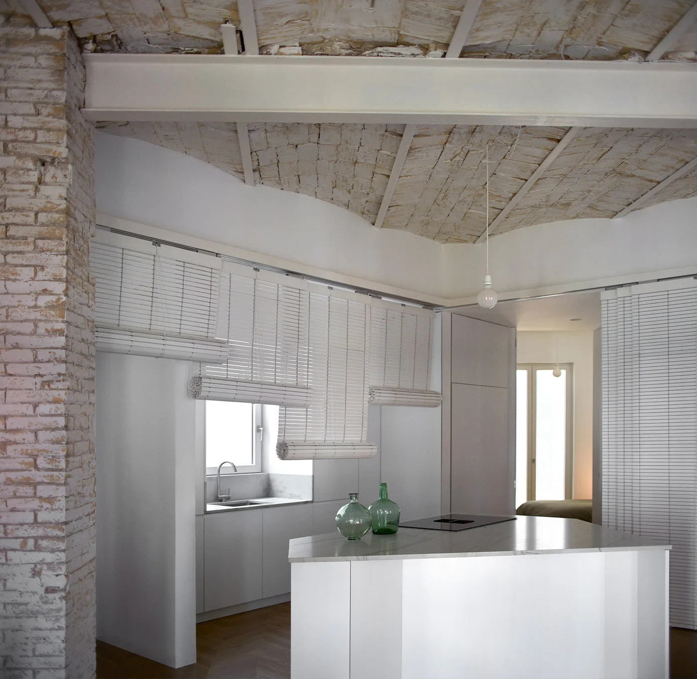 A minimalist white kitchen featuring a central island, modern flat-panel cabinets, and a rustic vaulted brick ceiling with a painted steel beam.