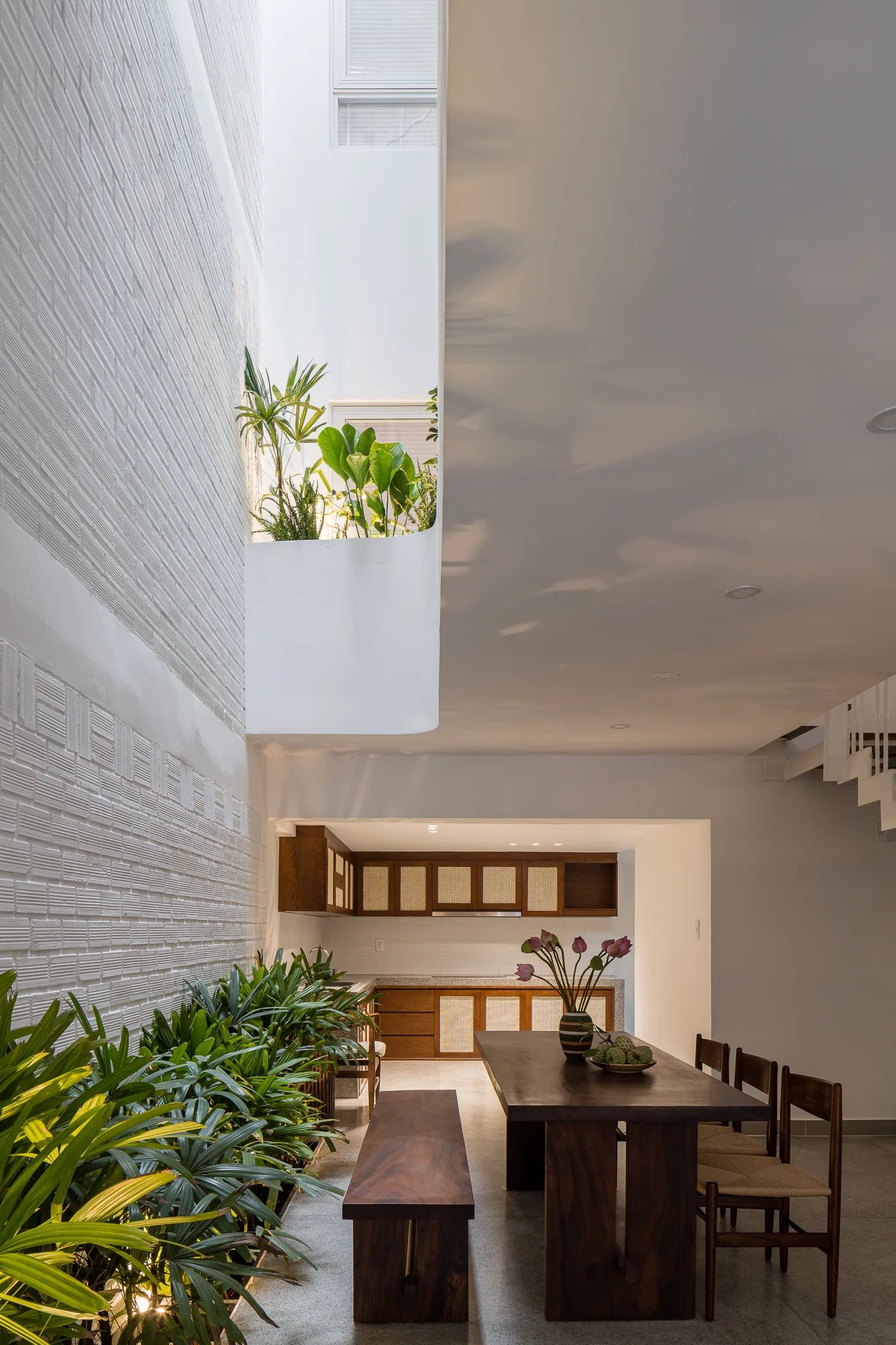 A modern dining room features a dark wood table and bench alongside a white brick wall lined with green indoor plants in an atrium.
