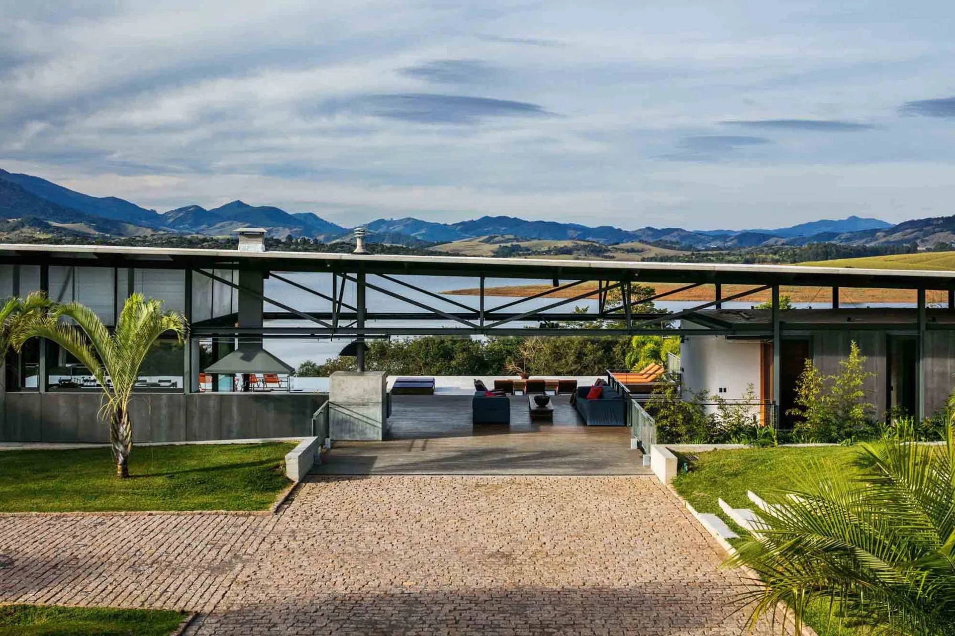 A modern open-sided deck with a steel truss roof and outdoor sofas overlooks a scenic lake and mountains at the end of a cobblestone path.