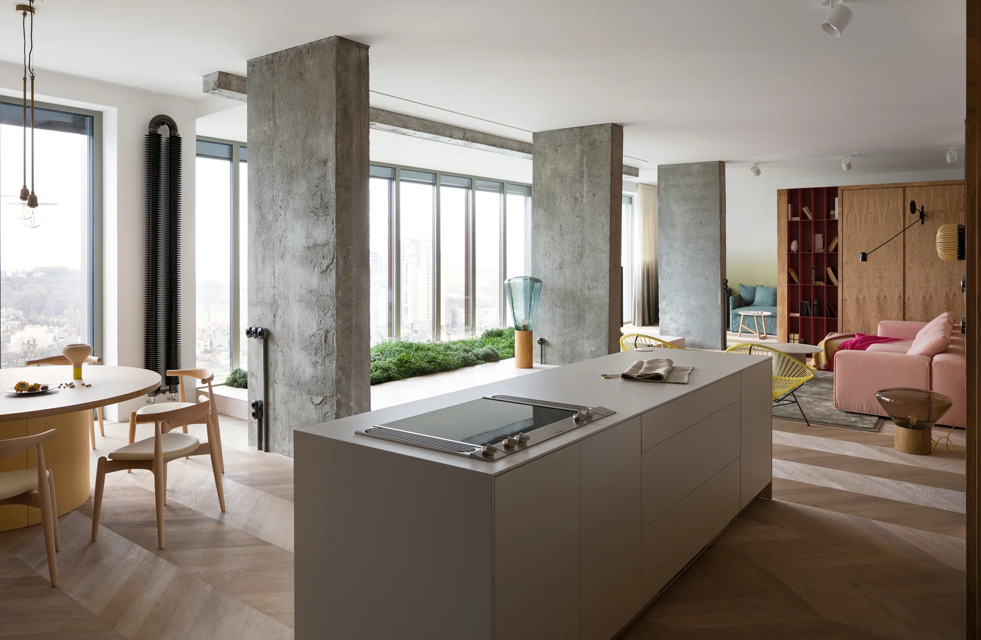 An open plan kitchen and living area featuring exposed concrete pillars, a chevron wood floor, a central kitchen island, and a dining table.