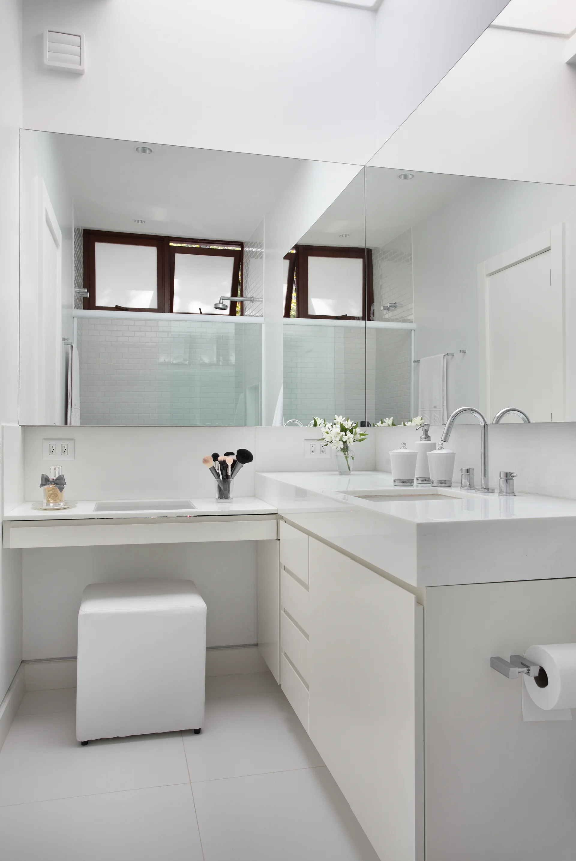 A minimalist white bathroom features a stepped vanity with a built-in makeup desk, a large wall mirror, and a square white stool.