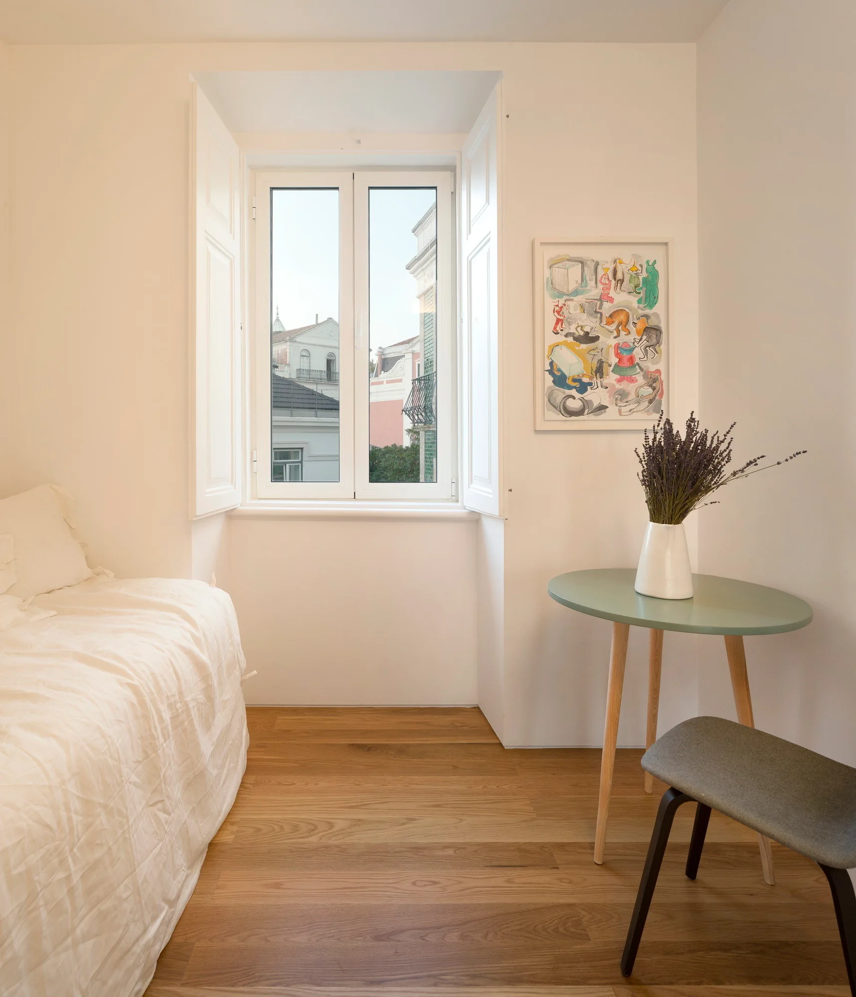 A minimalist bedroom featuring white bedding, a central window with open white shutters, and a small round green side table with a vase of lavender.