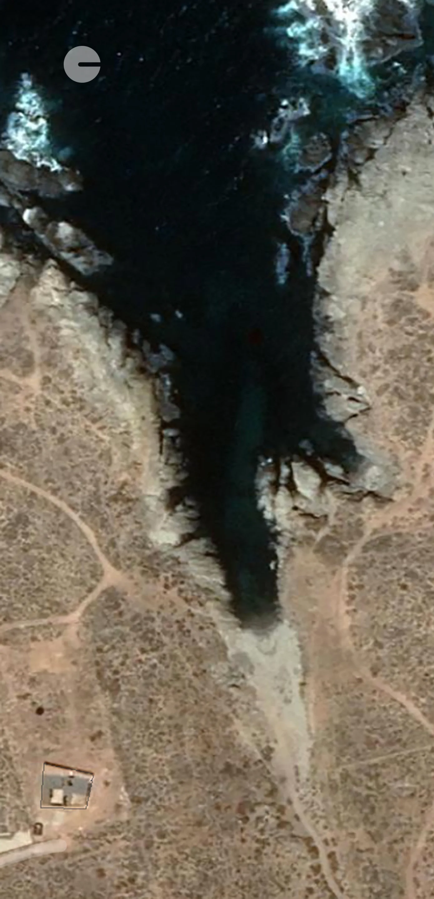 An aerial satellite perspective of a dark blue ocean inlet cutting into a rocky, tan arid landscape with faint trails.