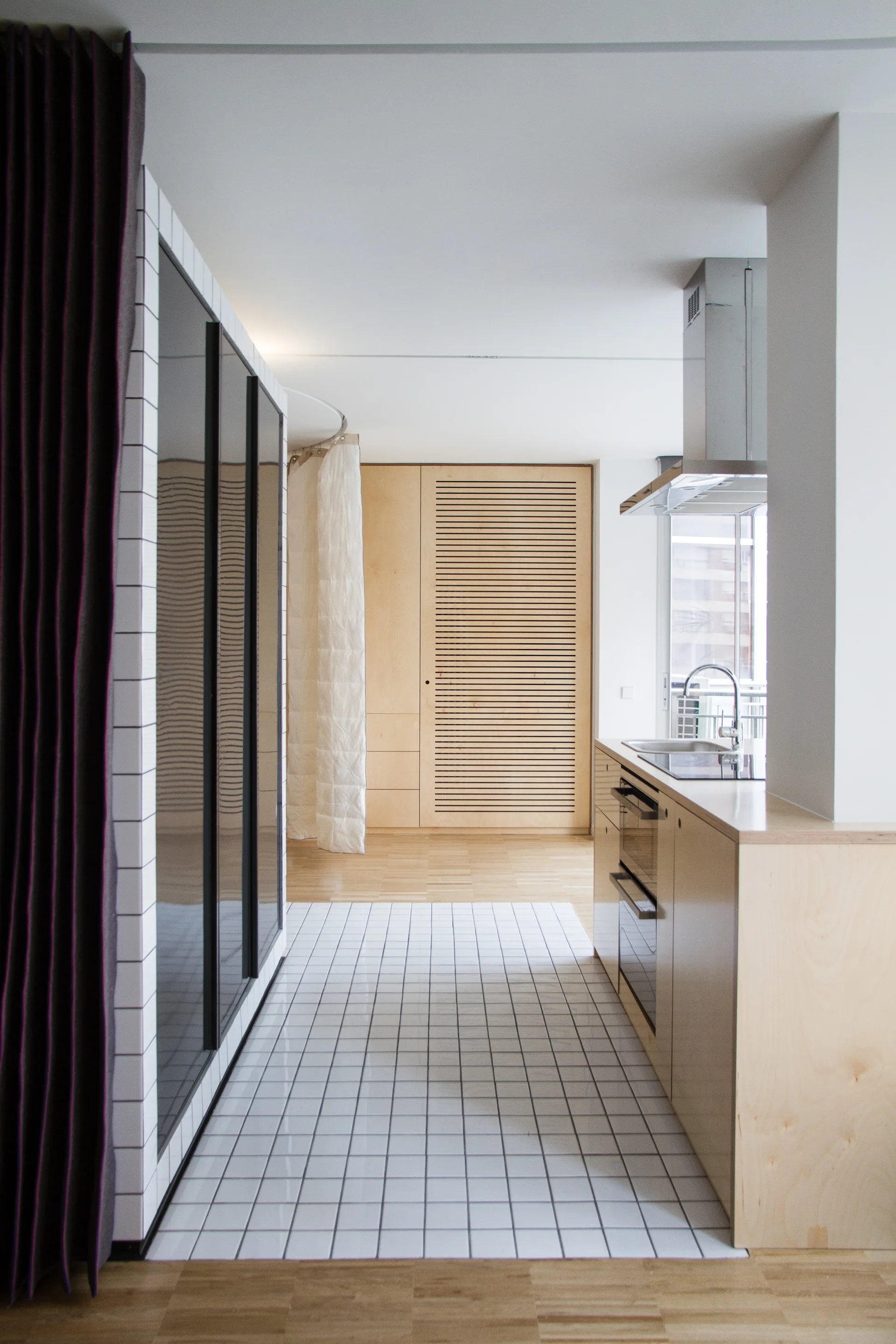 A minimalist kitchen features light wood cabinetry, a white square-tiled floor insert, a stainless steel hood, and a slatted wood door.