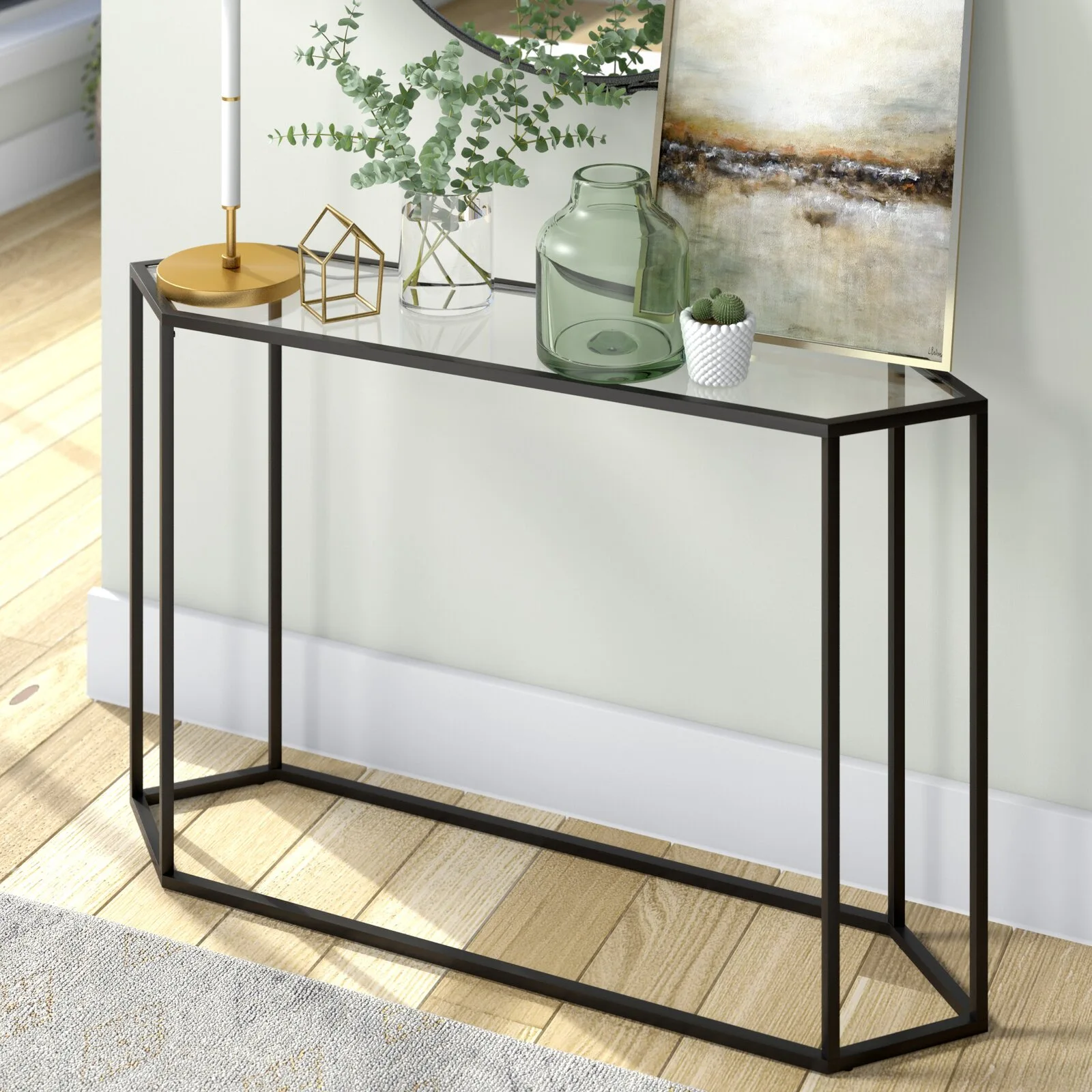 A narrow black metal console table with a glass top sits against a light wall on wood flooring, styled with vases and framed art.