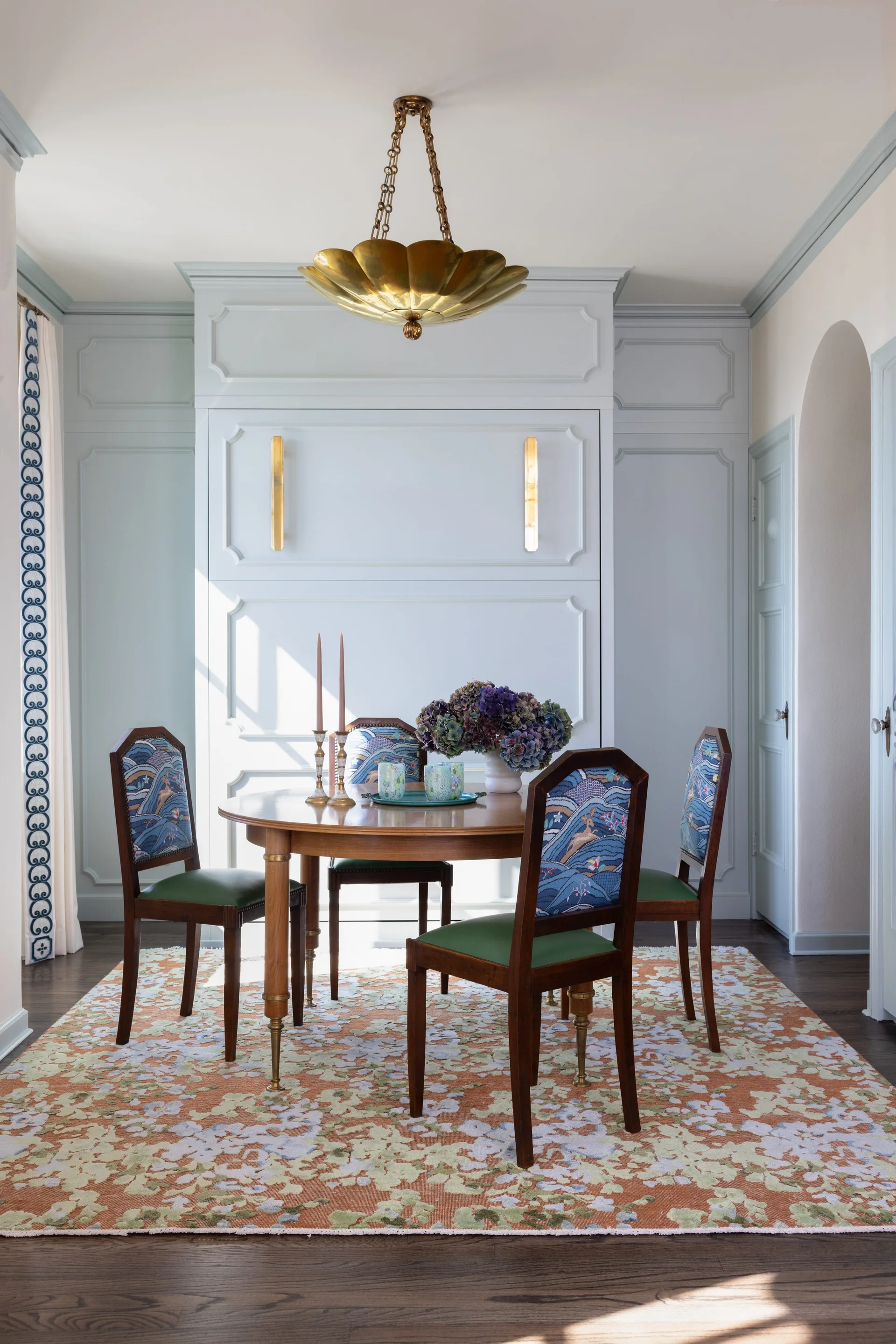 A round wood dining table and upholstered chairs with green seats are set against light blue paneled walls under a brass flower pendant light.