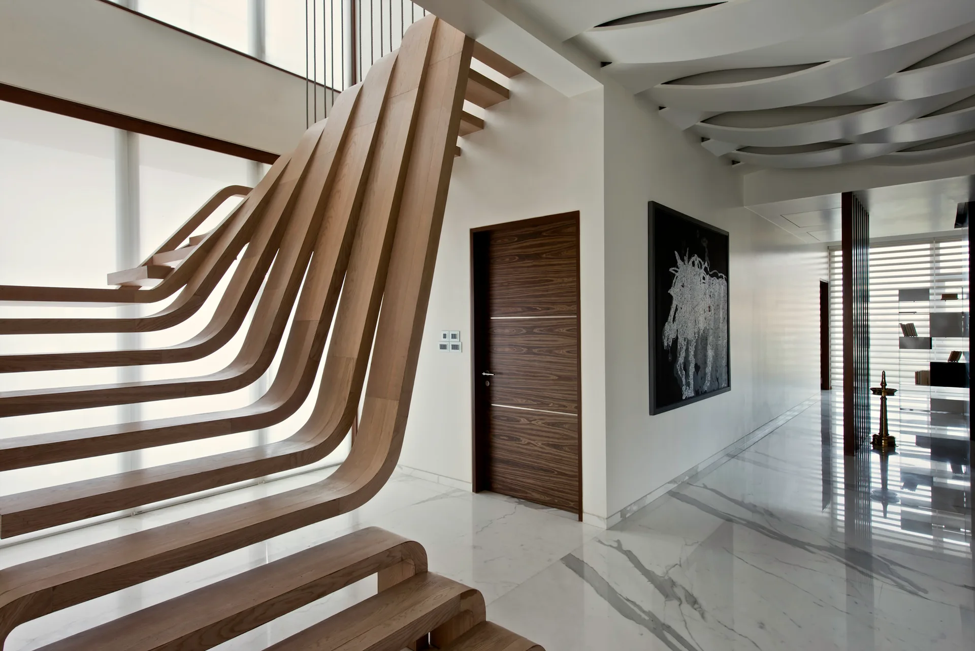 A modern hallway features a sculptural curved wood slat staircase, glossy white marble flooring, and a dark wood door next to abstract artwork.