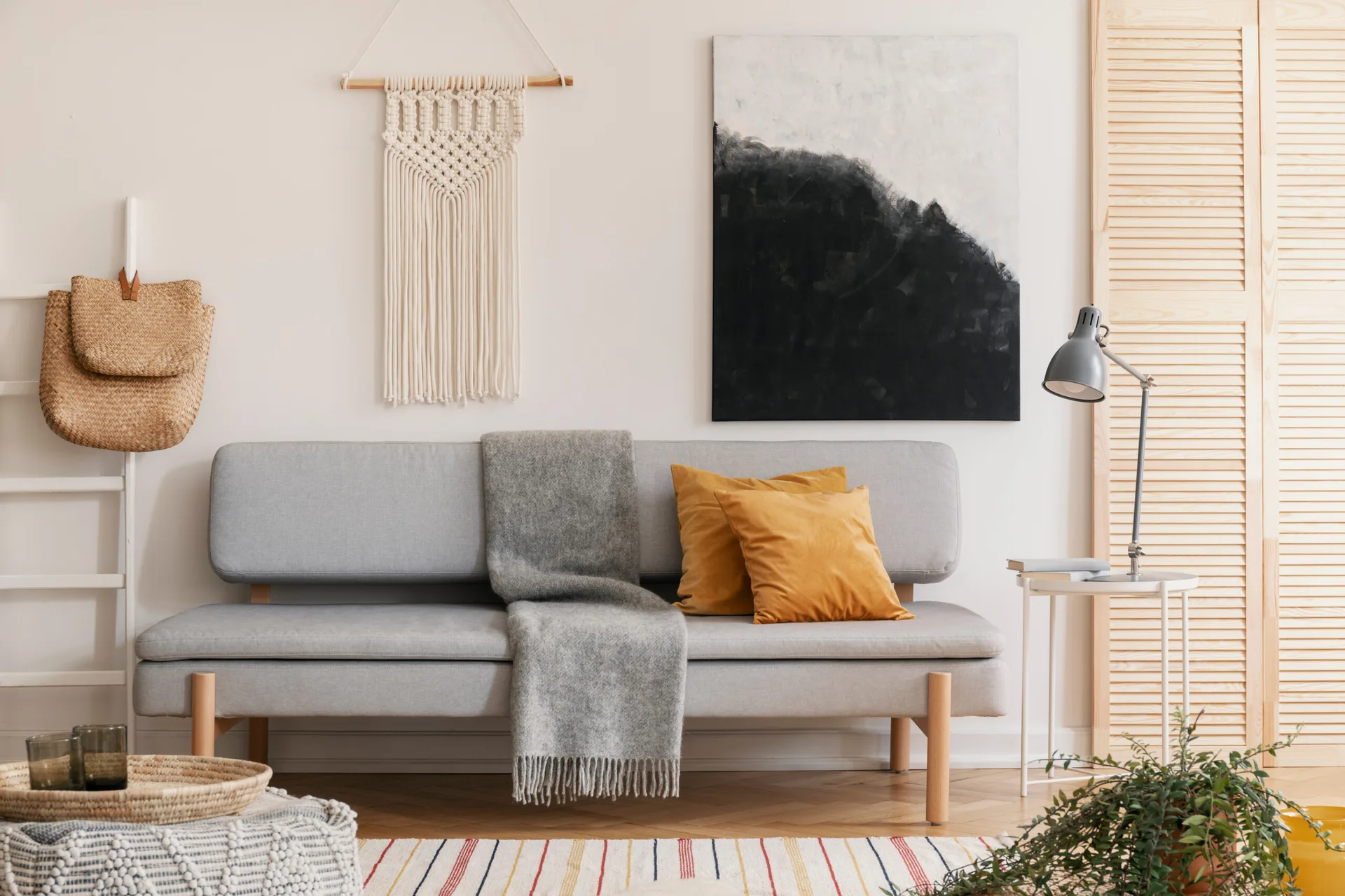 A light grey sofa with ochre yellow pillows is set against a wall featuring an abstract black painting and a white macrame hanging.