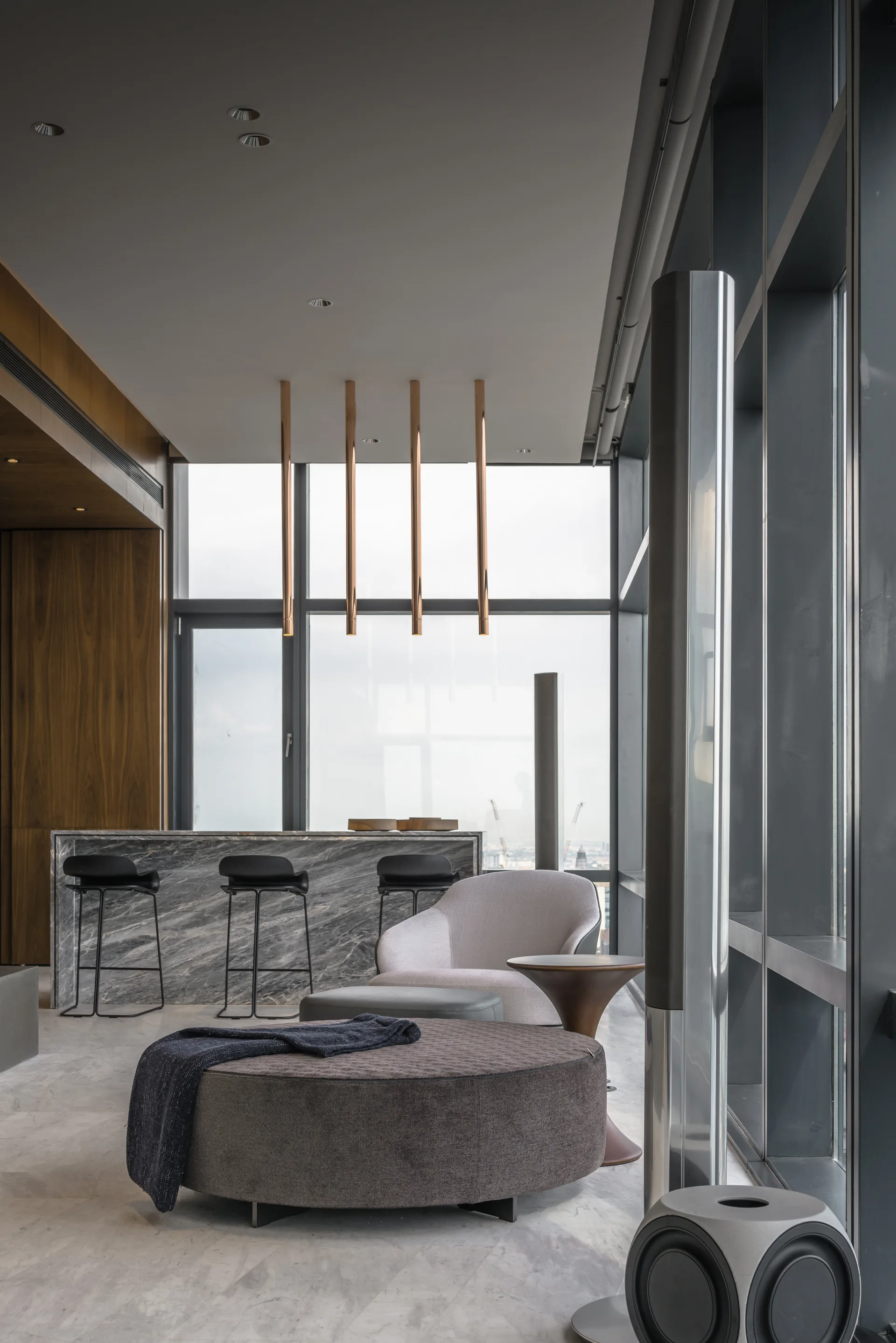 A modern lounge area with a round grey ottoman and a lounge chair near a marble kitchen bar with black stools and copper pendant lights.