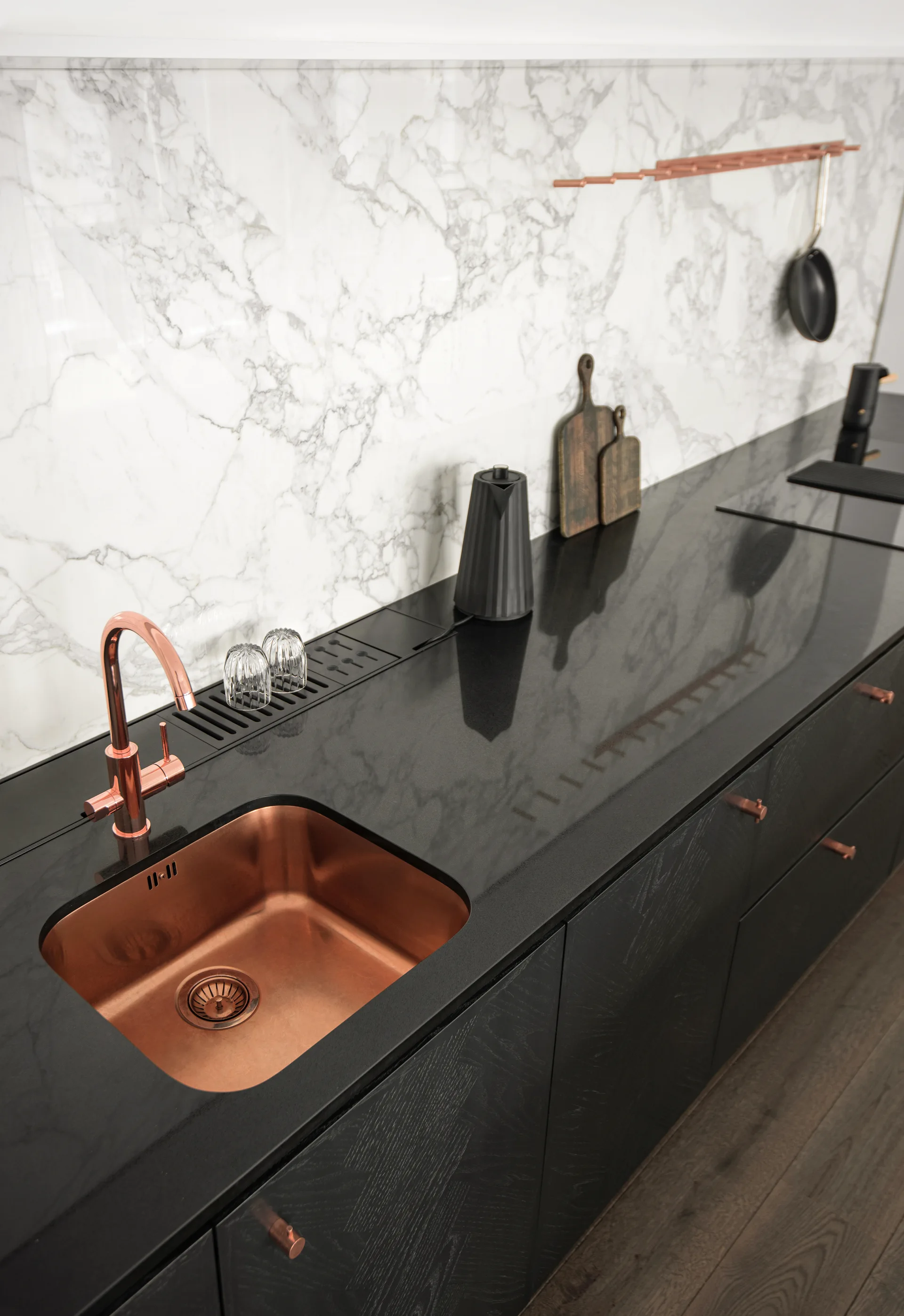 A copper undermount sink and faucet are set in a black countertop against a heavily veined white marble backsplash.