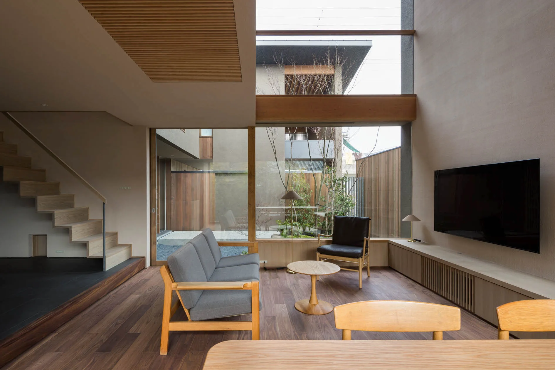 A modern living room features a gray sofa, wood-plank floors, a floating staircase, and a large glass wall looking out to a courtyard.