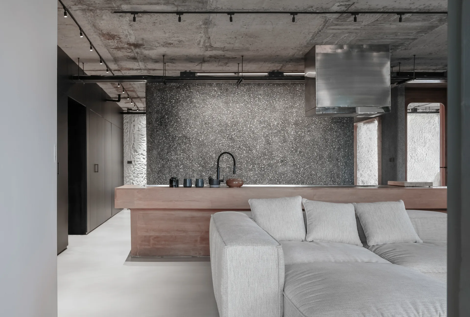 A modern living room features a beige sofa in front of a wooden kitchen island, backed by a grey speckled terrazzo wall and a raw concrete ceiling.