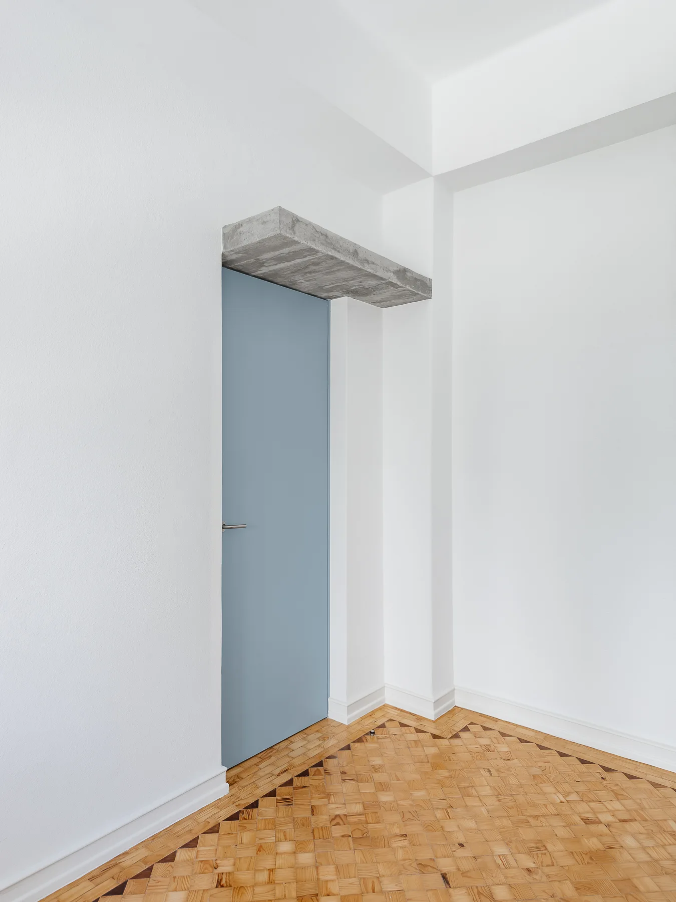 A light blue minimalist interior door topped by a raw concrete block lintel against white walls and a patterned wood parquet floor.