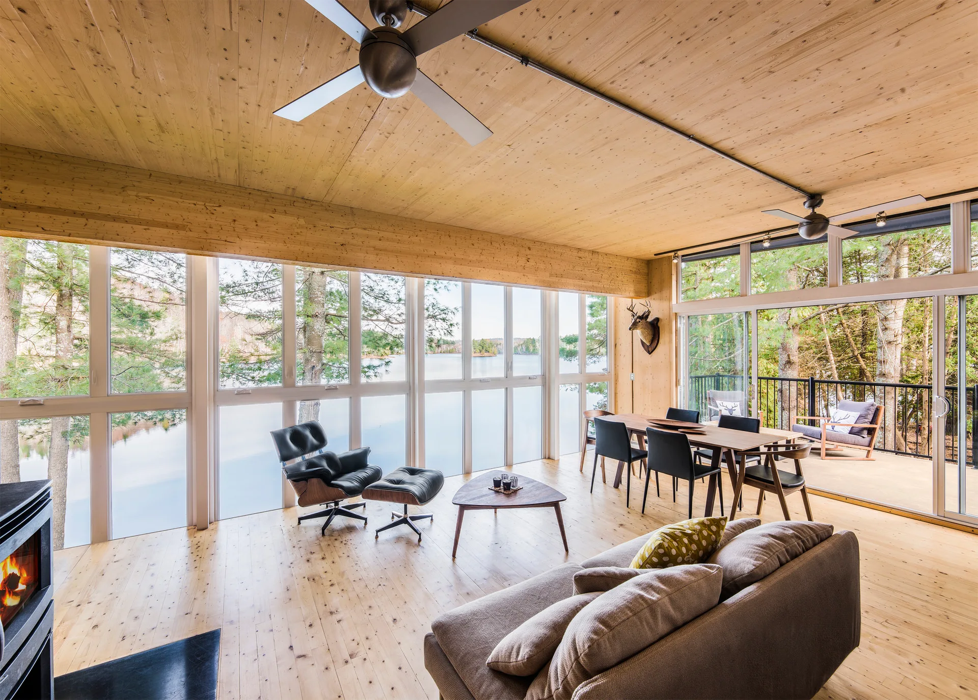 An open-concept cabin living room featuring a black lounge chair, a dining area, and floor-to-ceiling windows showing a view of a lake.