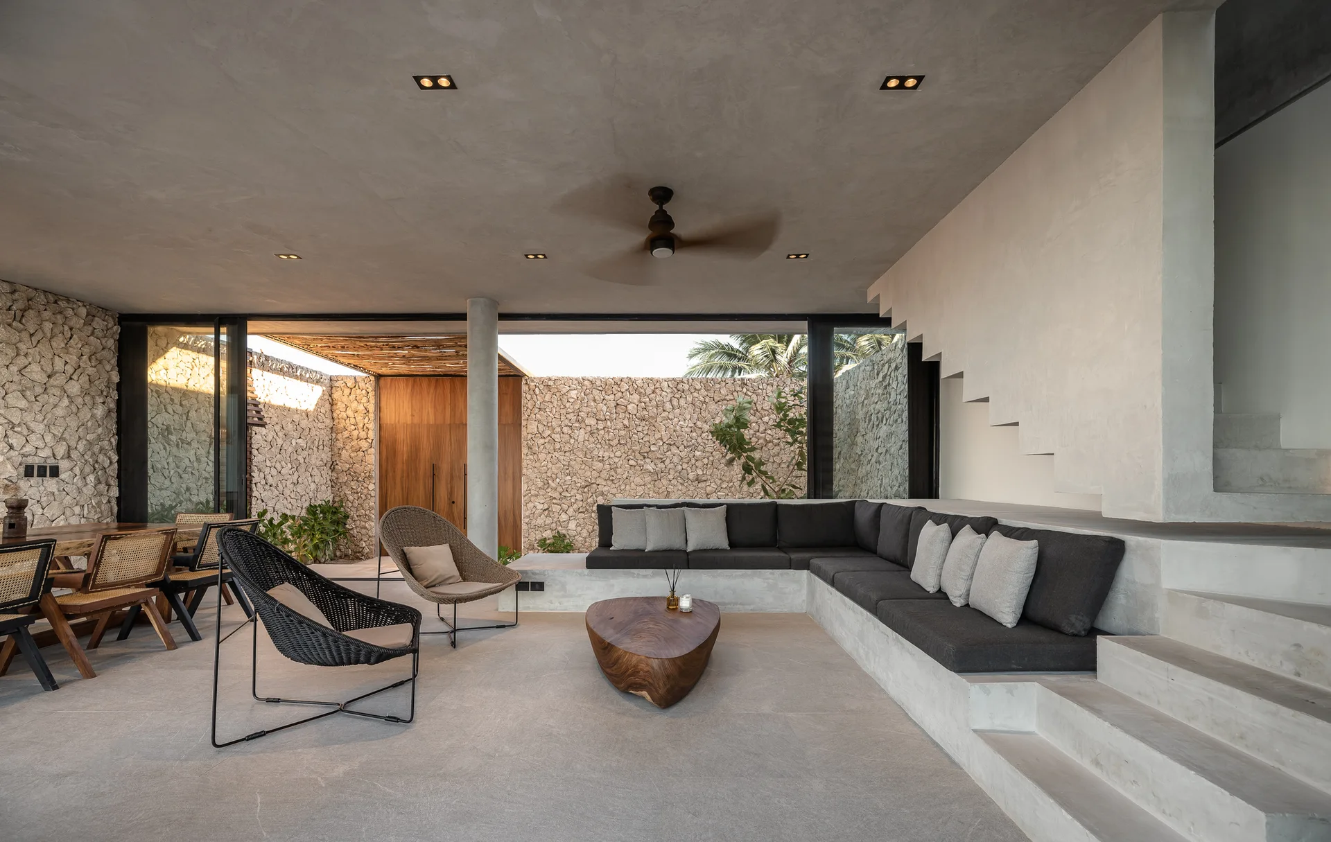 A covered concrete patio features a built-in grey sectional sofa, an organic wood coffee table, and a background of stacked stone walls.