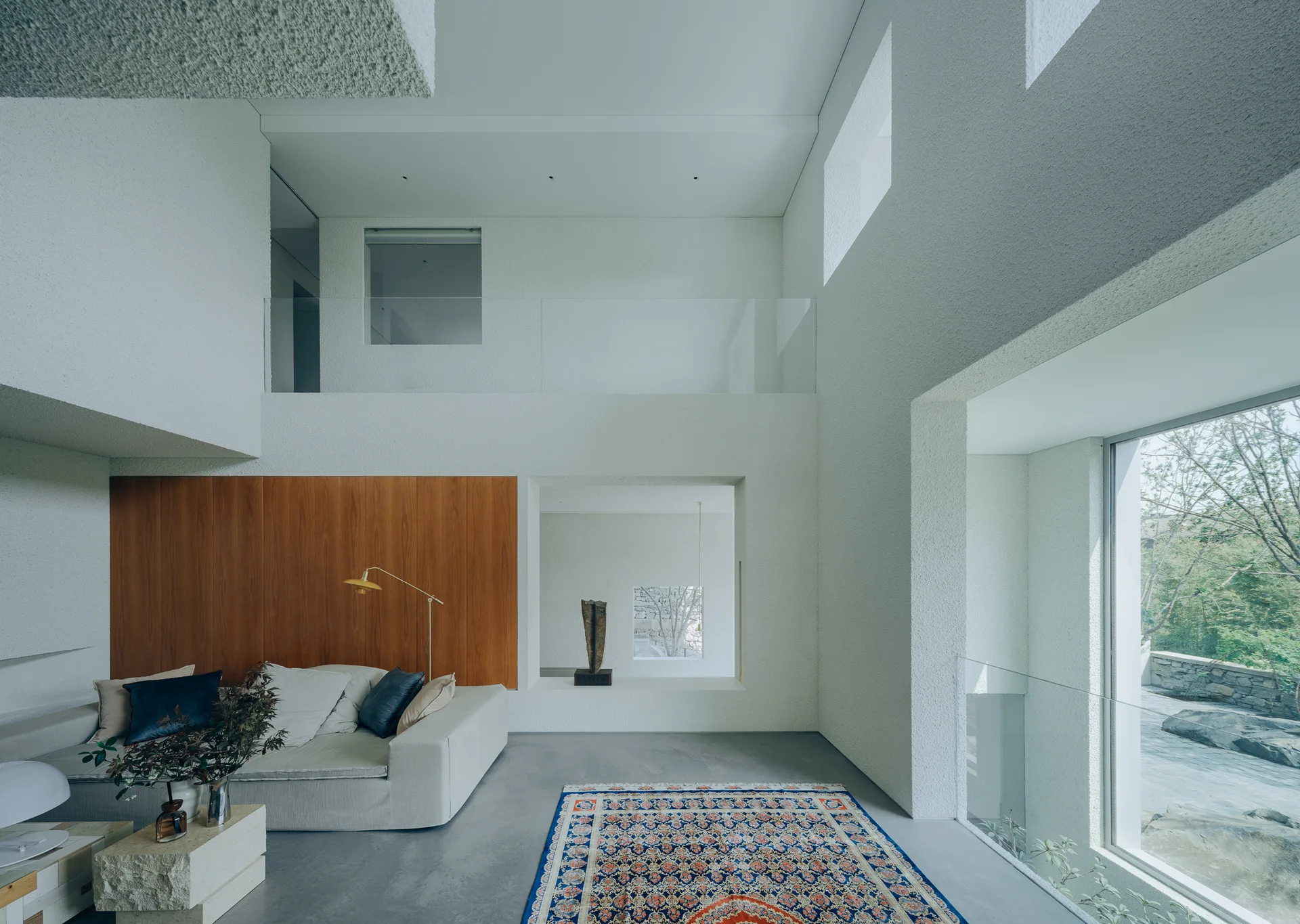 A double-height living room with white stucco walls, a wood accent panel, a cream sofa, and an ornate rug on polished concrete flooring.