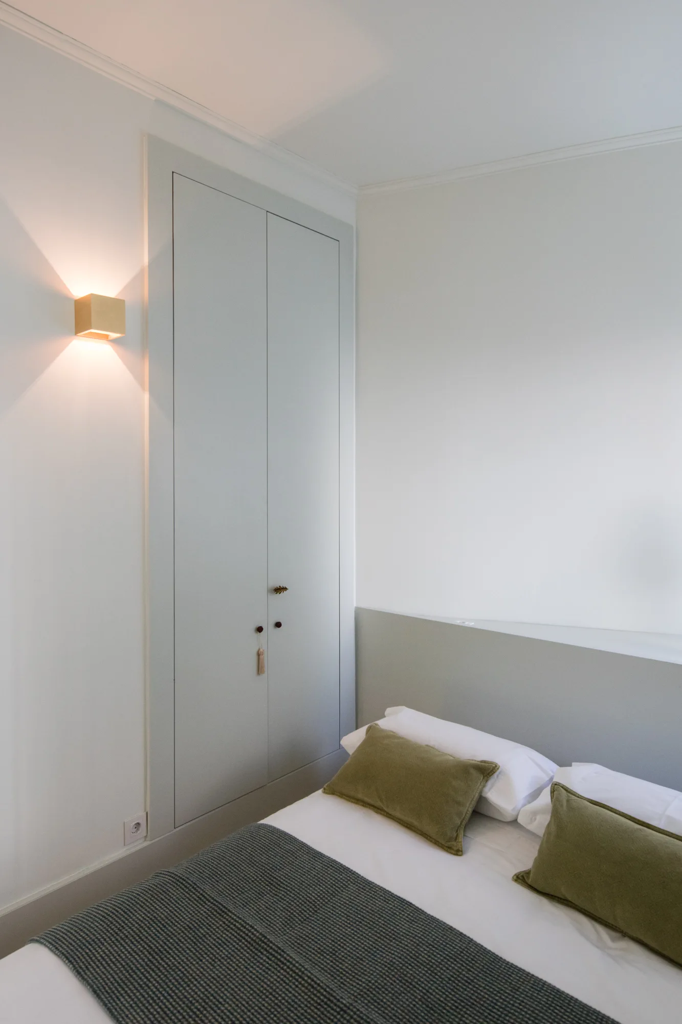 A minimalist bedroom featuring a bed with olive green pillows, a low grey headboard ledge, a built-in grey wardrobe, and a glowing cube wall sconce.