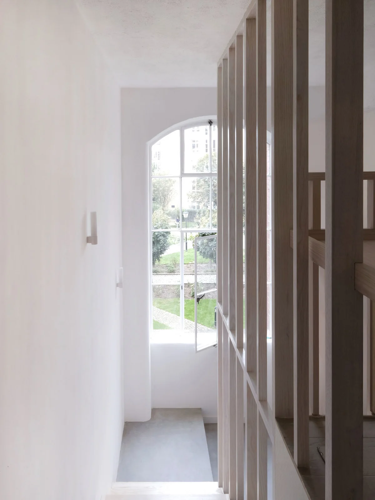 A minimalist white staircase features a vertical light wood slatted screen and an arched window looking out onto a green garden.