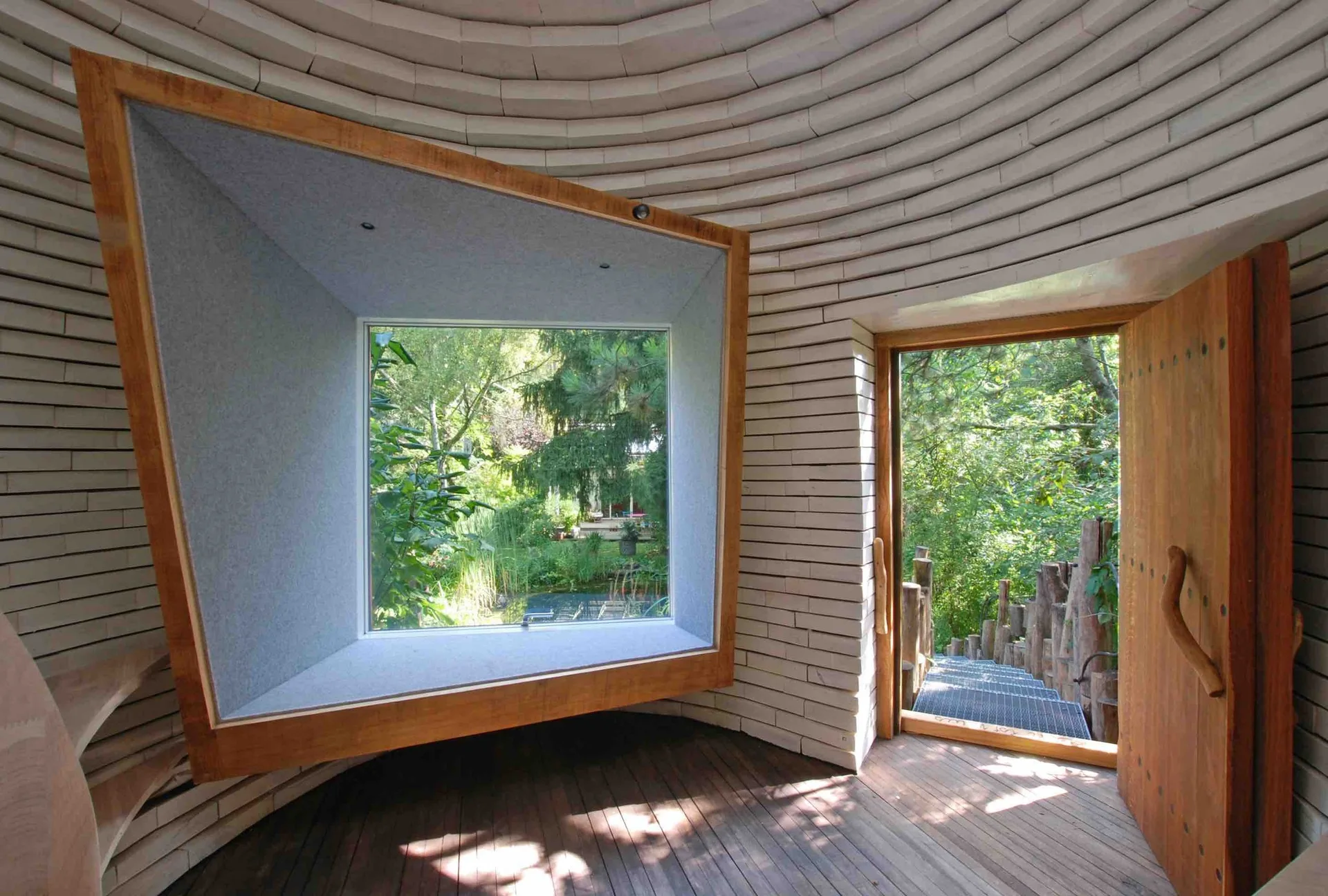 A circular room with white stacked brick walls features a large rectangular window framing a green garden and an open wooden door.