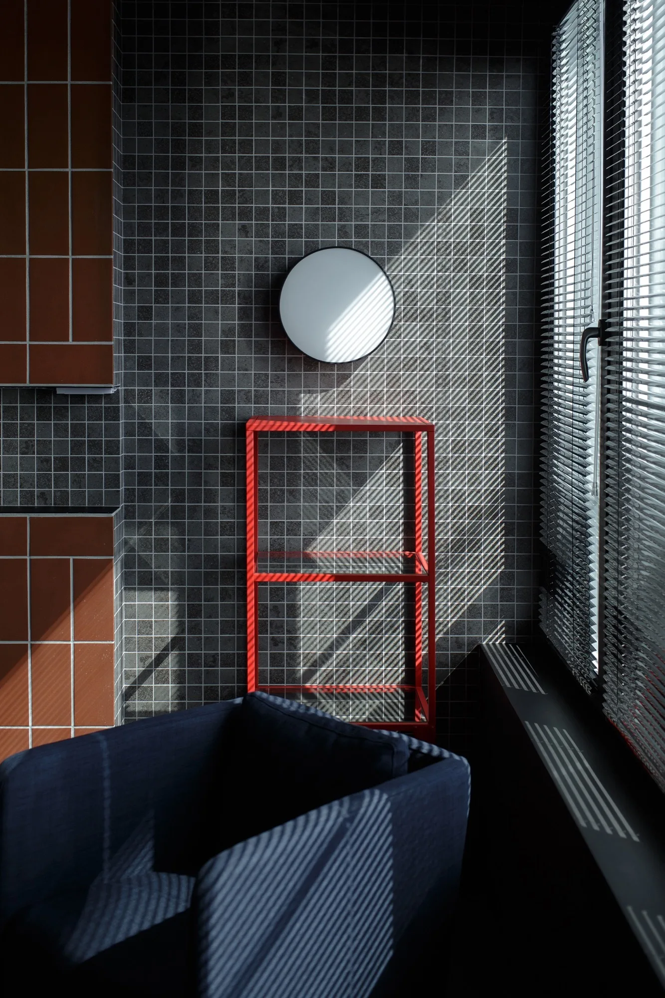 A navy blue armchair sits before a gray tiled wall with a bright red metal shelf, a circular mirror, and window blinds.