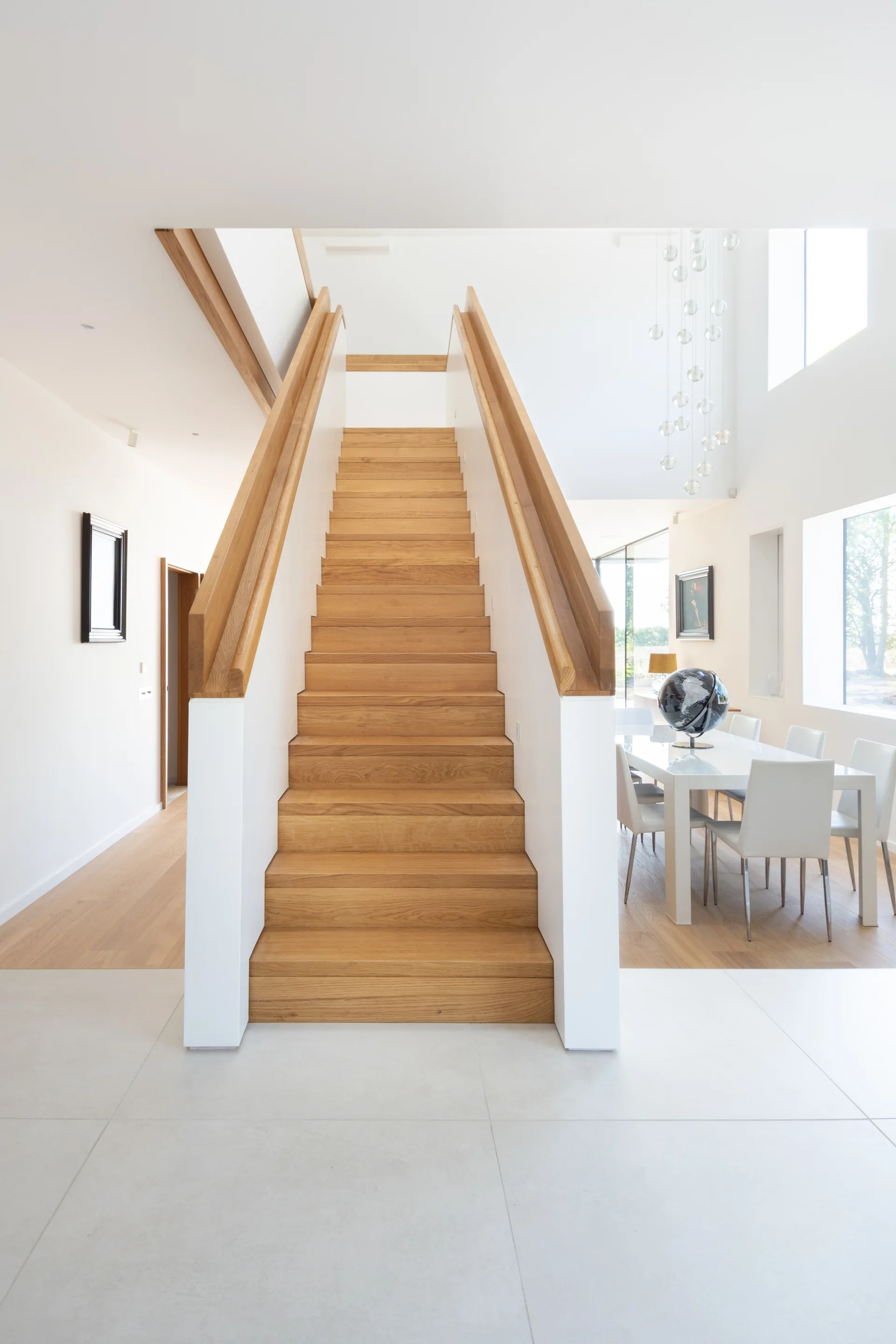 Central modern wooden staircase with white side walls leading upstairs next to a bright open-plan dining room with white chairs.
