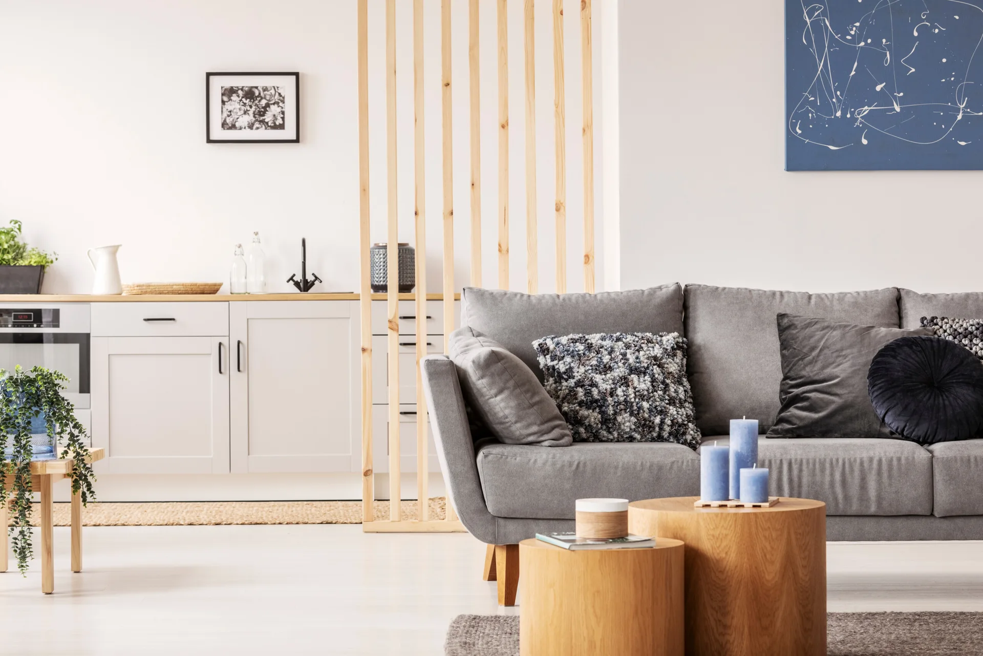 A living room features a grey fabric sofa, two round wooden coffee tables, and a vertical wood slat screen separating the space from a white kitchen.