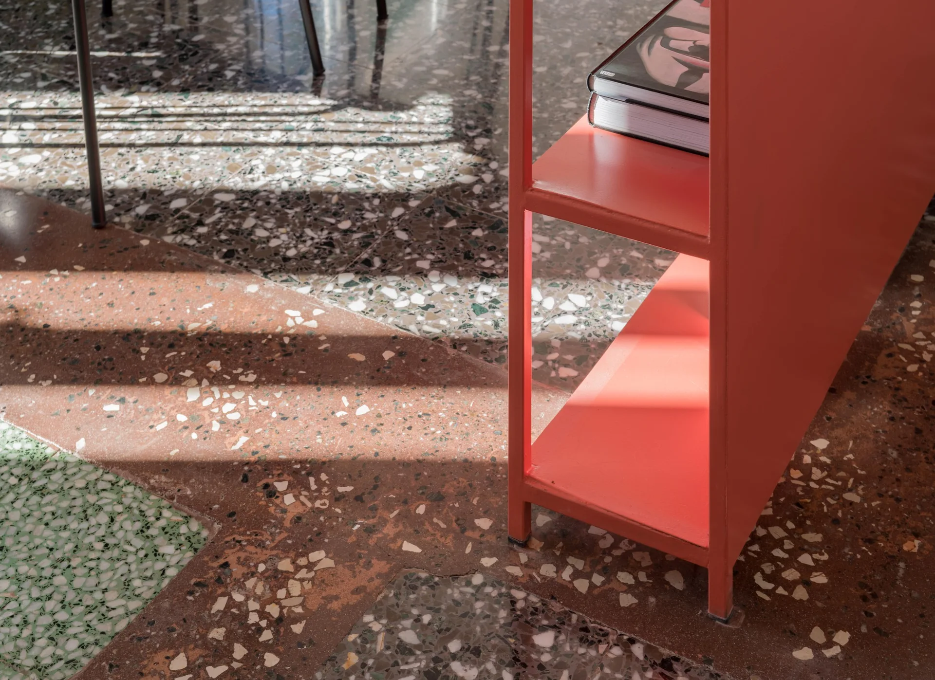 A bright coral metal shelving unit holding books sits on a polished, multi-colored terrazzo floor with diagonal sunlight shadows.